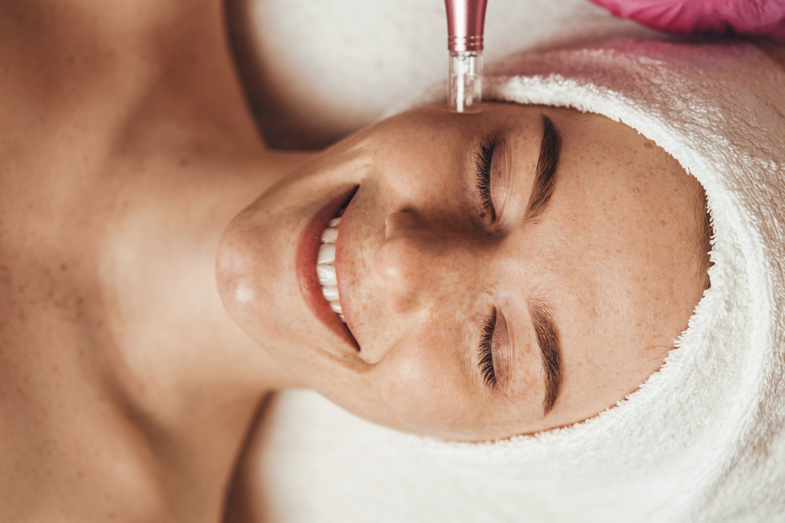 A woman receives a facial treatment at a spa, with her eyes closed and covered with a towel, while a professional applies a facial device on her face.
