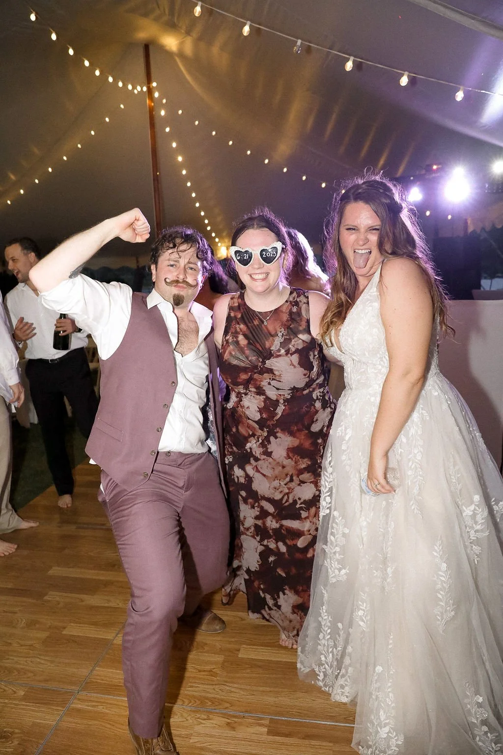 Three people at a wedding reception, with the man on the left flexing his arm and the woman on the right sticking out her tongue. The woman in the middle is wearing themed sunglasses with the date '6-21-2025'. The background shows string lights and o