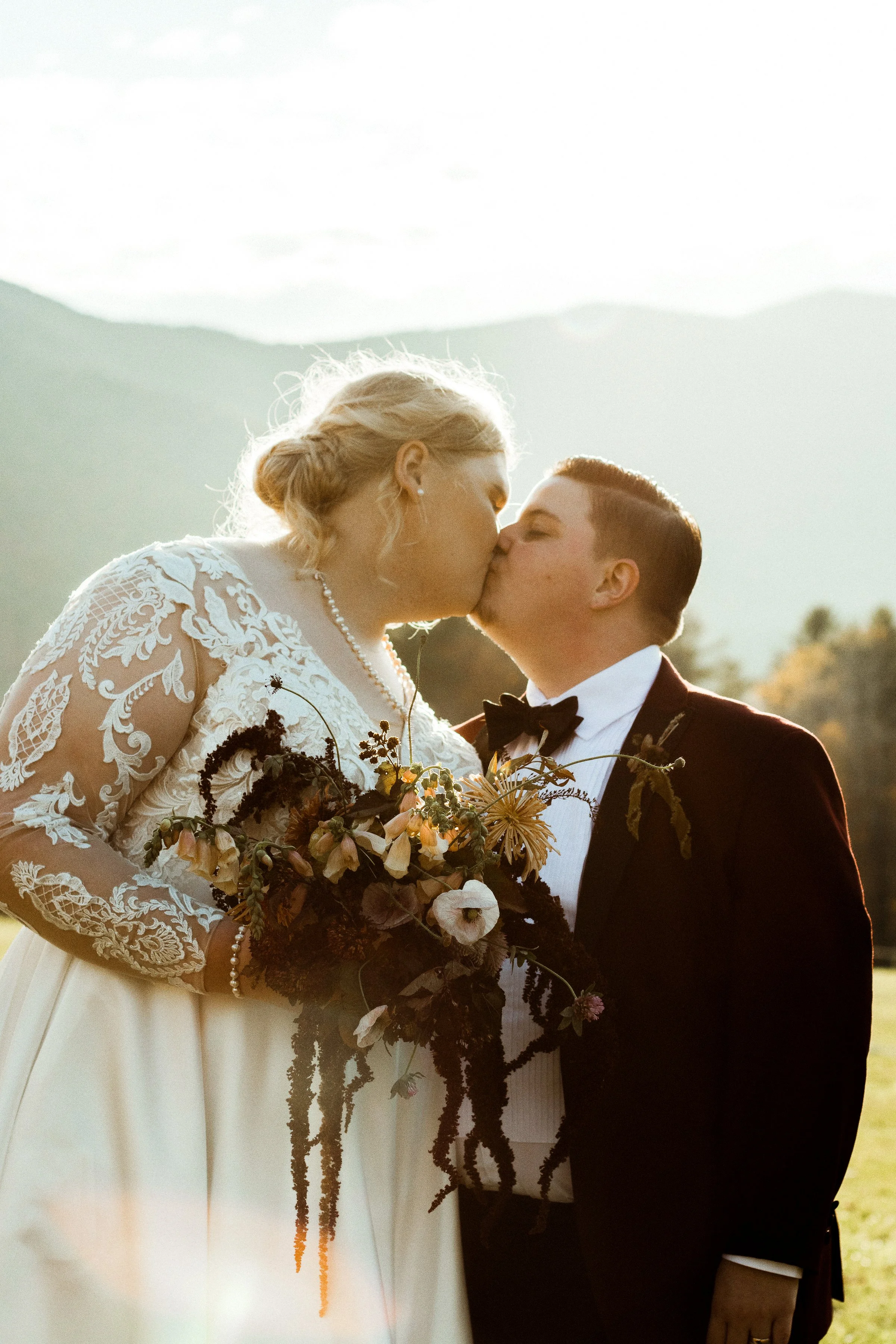 A bride and groom kiss outdoors during sunset, with mountains and a cloudy sky in the background. The bride wears a white lace wedding dress and holds a floral bouquet, while the groom wears a tuxedo with a bow tie.