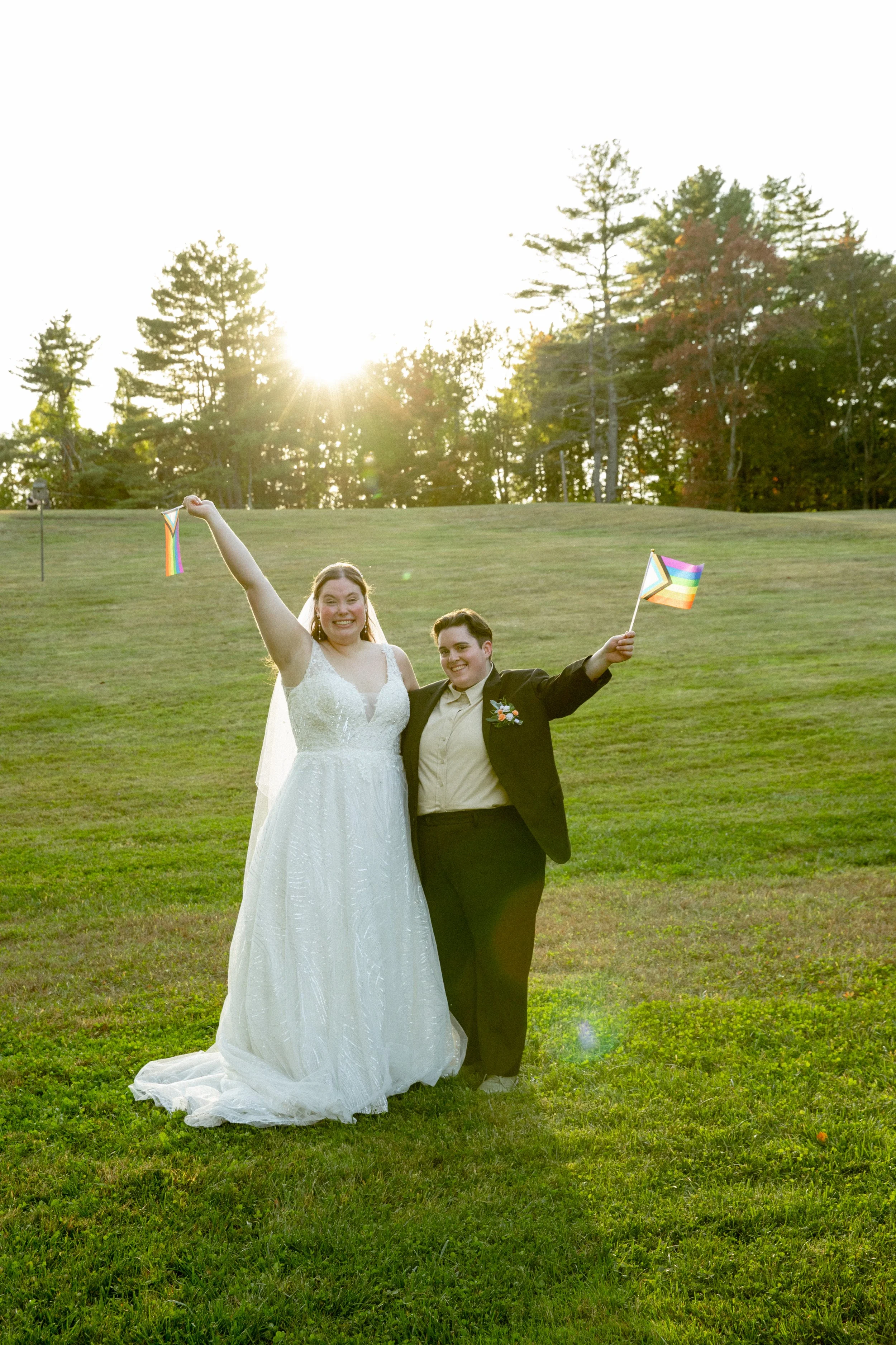 Two people celebrating LGBTQ+ pride at a wedding, one wearing a white wedding dress and the other in a black suit, holding LGBTQ+ pride flags in an outdoor setting during sunset.