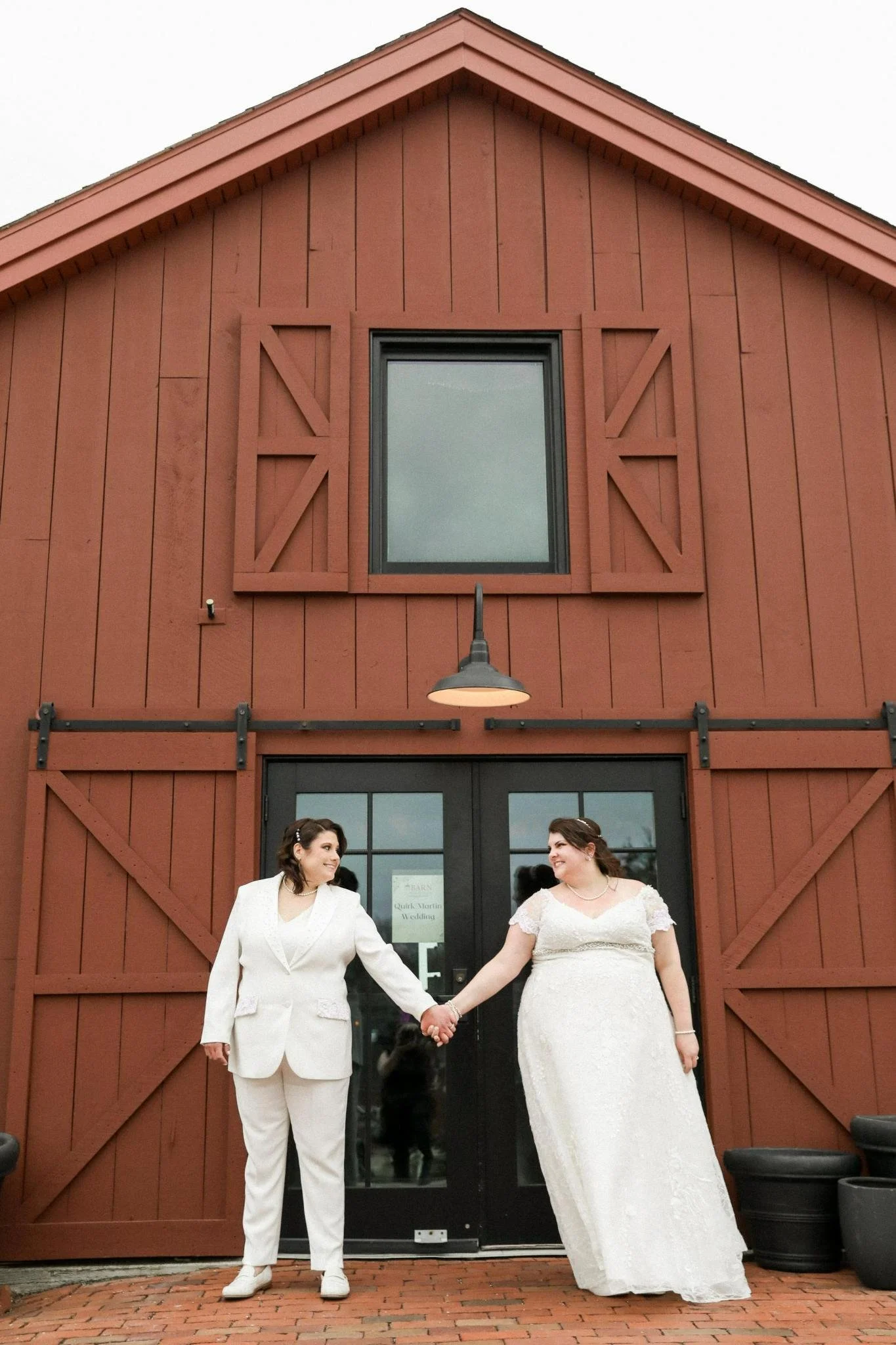 Two women in wedding dresses holding hands in front of a barn with red siding and black door.