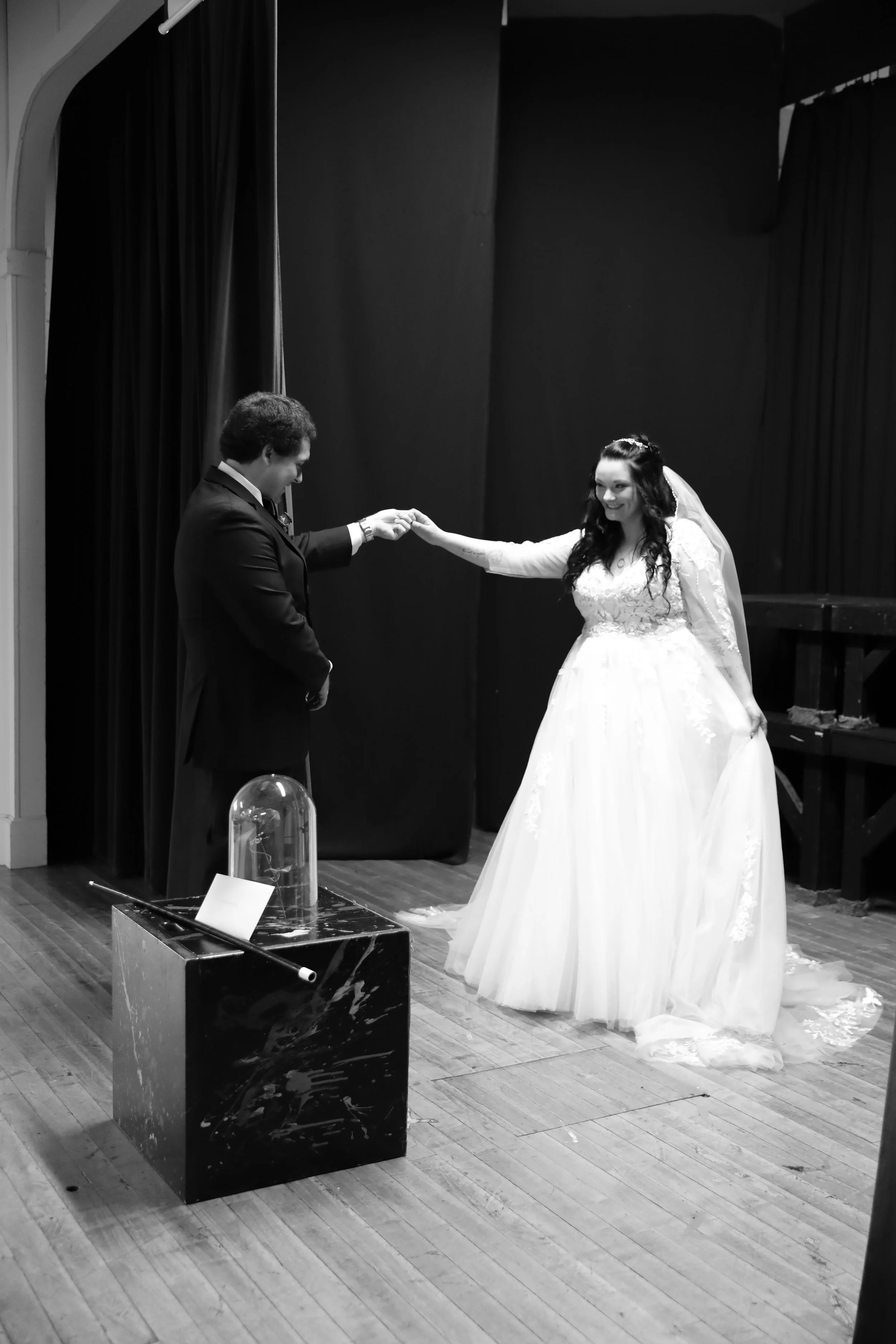 A black-and-white photo of a wedding ceremony where a bride and groom are holding hands. The bride is smiling and wearing a long-sleeved lace wedding dress with a veil. The groom, dressed in a suit, is looking at her. A glass container and a sign are on a table in front of them, and the scene is set against a dark curtain background.