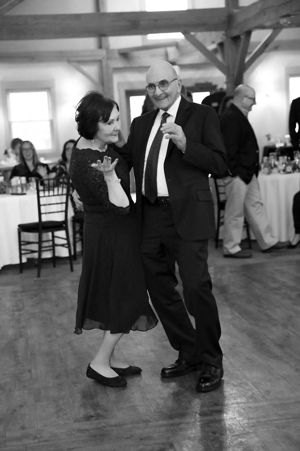 A black and white photo of a man and woman dancing closely at a formal event. The woman is wearing a dark dress and flats, the man is wearing a suit, tie, and glasses. In the background, there are other guests seated at tables and standing, with wood