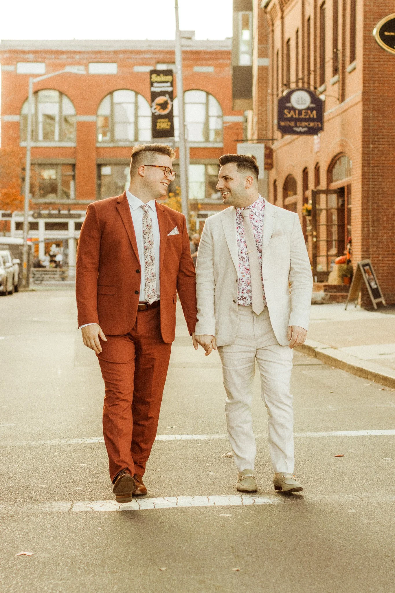 Two men dressed in suits holding hands walking on a city street, smiling at each other.