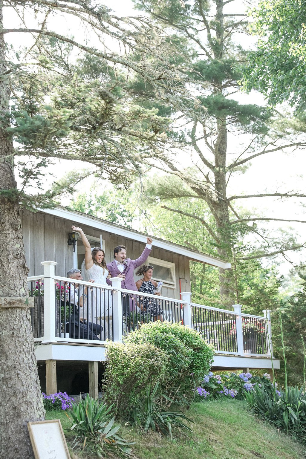 A group of six people celebrating on a deck outdoors surrounded by trees and greenery. They are dressed in formal attire, with some raising their arms and smiling.