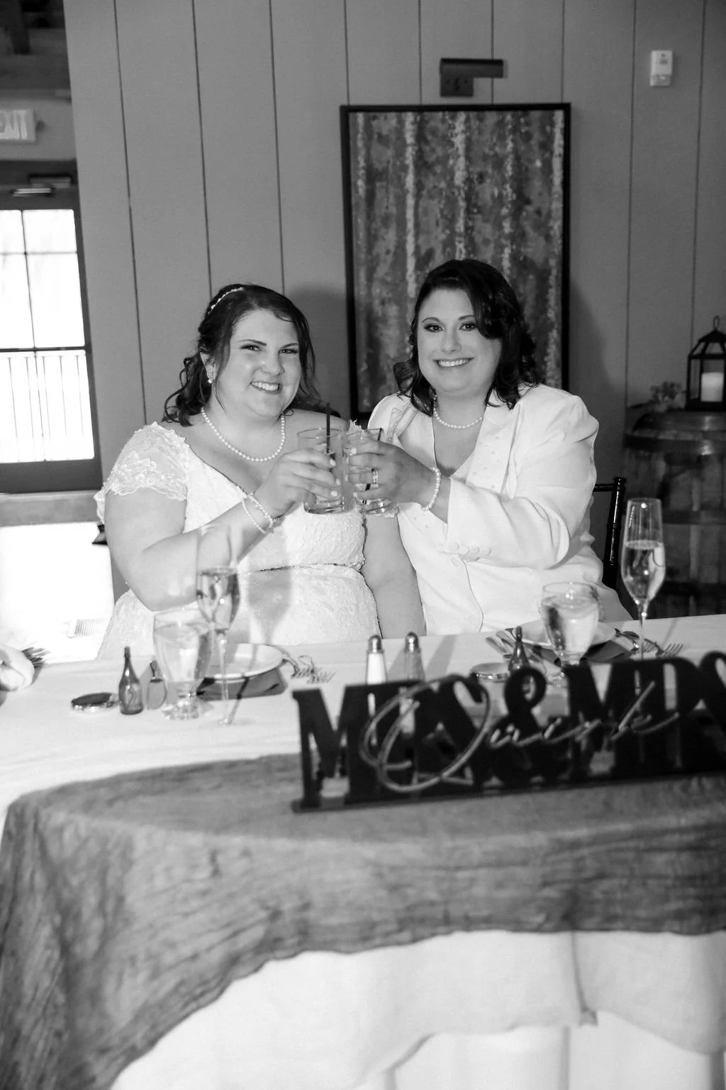 Two women, one in a wedding dress and the other in a white blazer, smiling and holding glasses in a toast at a wedding reception.