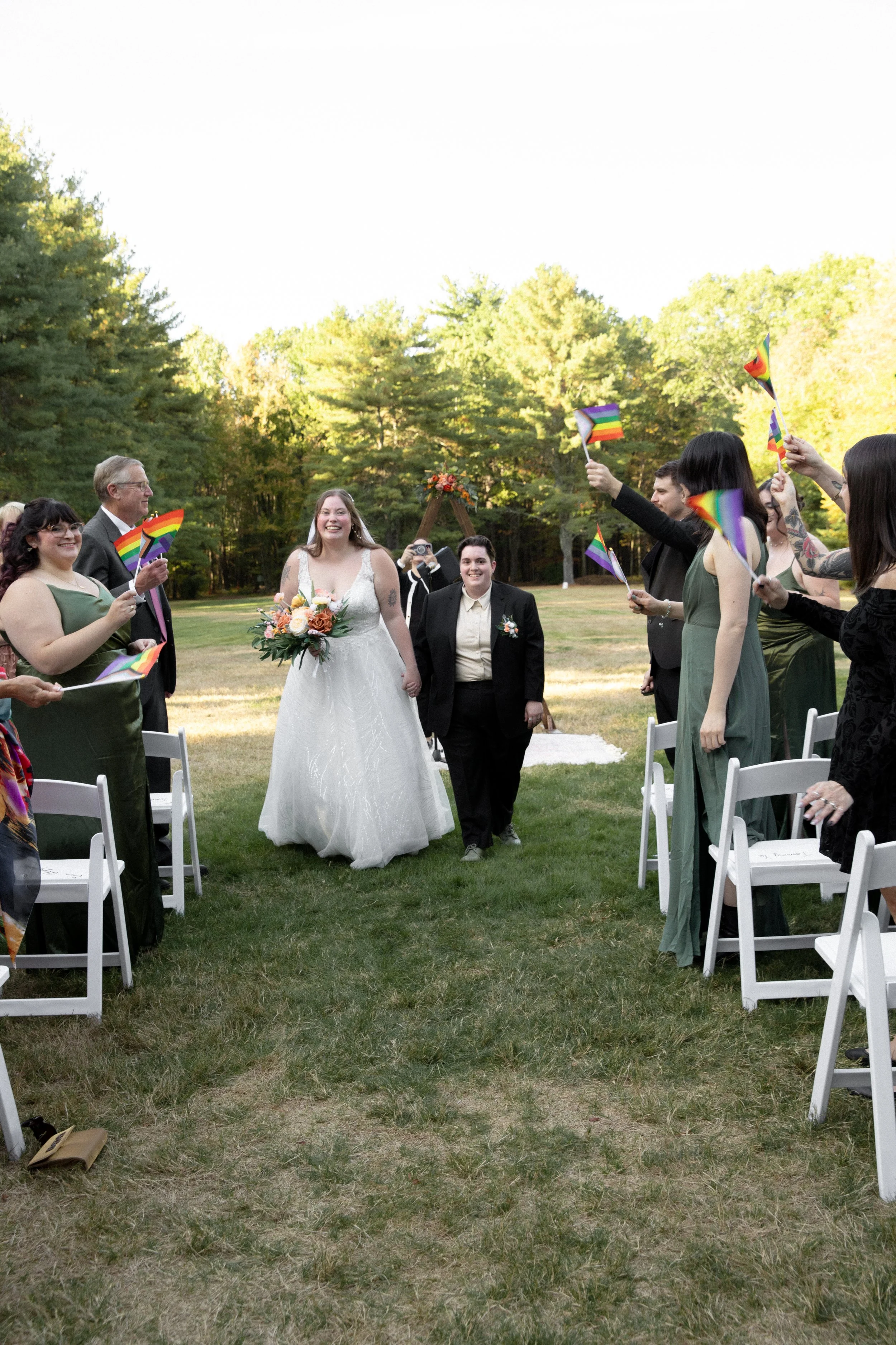 Bride and groom walking down the aisle at their outdoor wedding ceremony surrounded by friends and family waving rainbow pride flags.