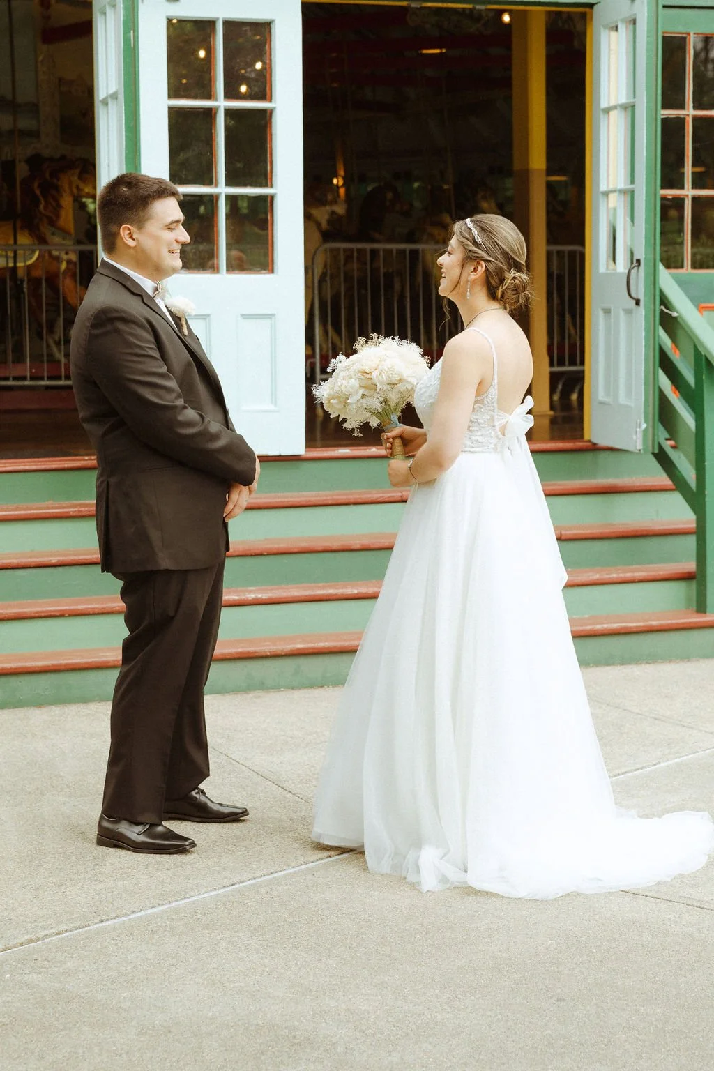 Bride in a white wedding dress holding a bouquet of flowers, facing groom in a black suit, outside a building with open doors, stairs, and green railings, during wedding ceremony.