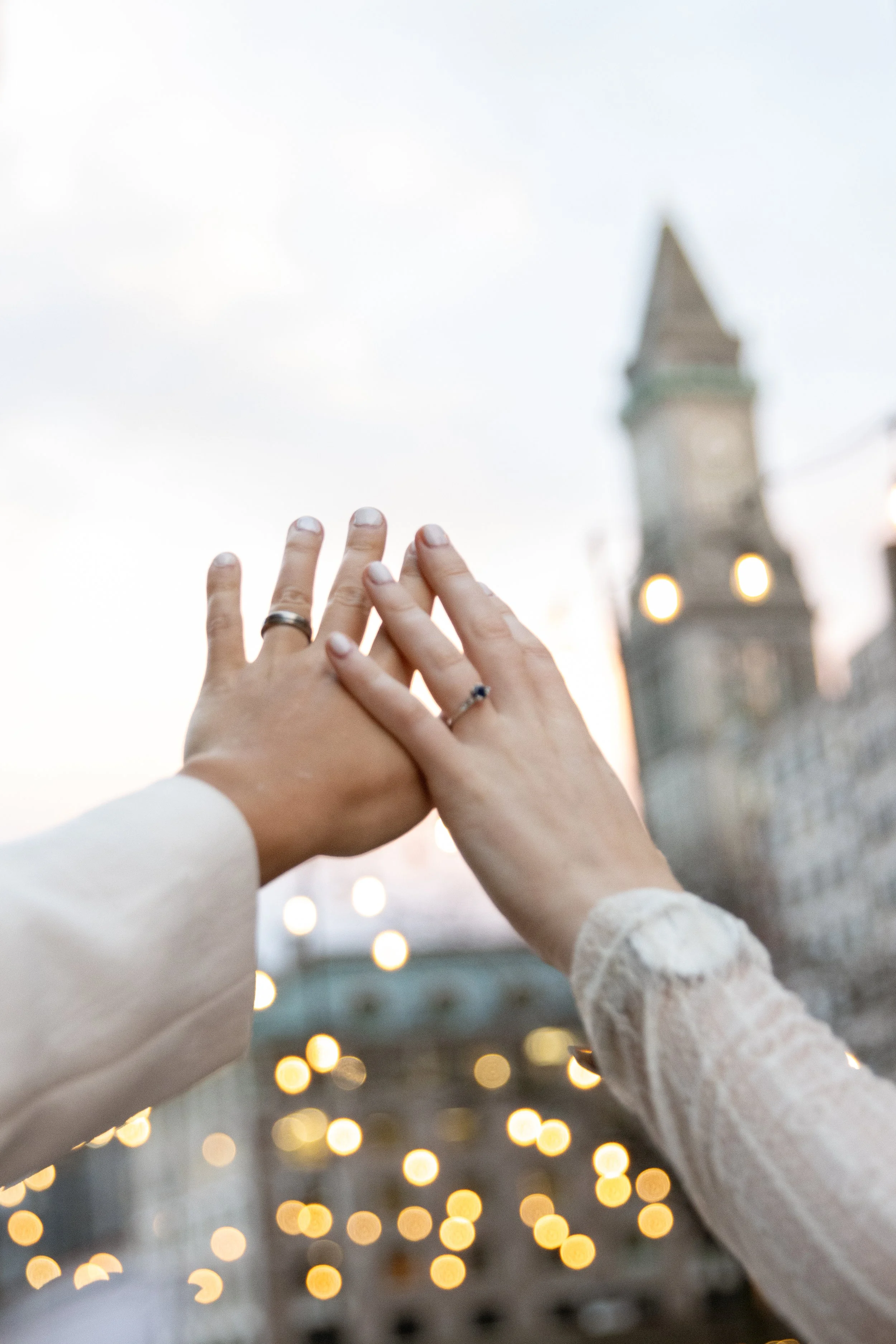 Two hands, each with a wedding ring, touching with a blurred historic building and lights in the background.