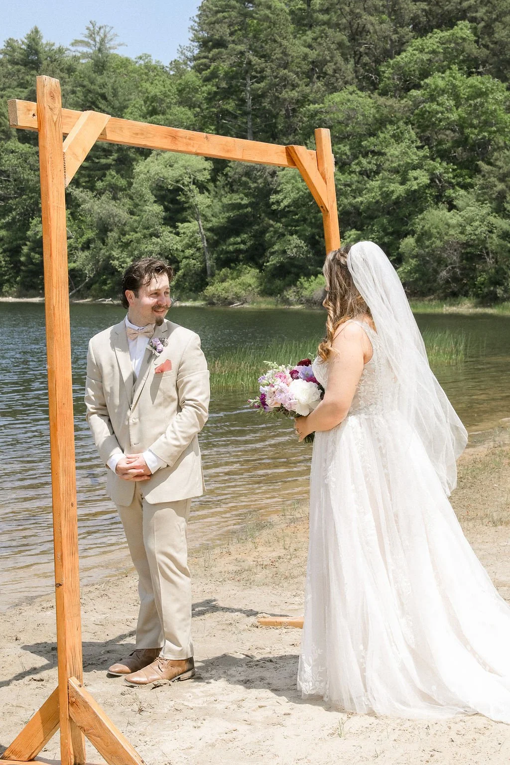 A bride and groom standing on a sandy beach by a lake during a wedding ceremony. The groom is wearing a light beige suit with a bow tie, and the bride is in a white wedding dress with a veil holding a bouquet of flowers. They are under a wooden arch, surrounded by green trees and water.