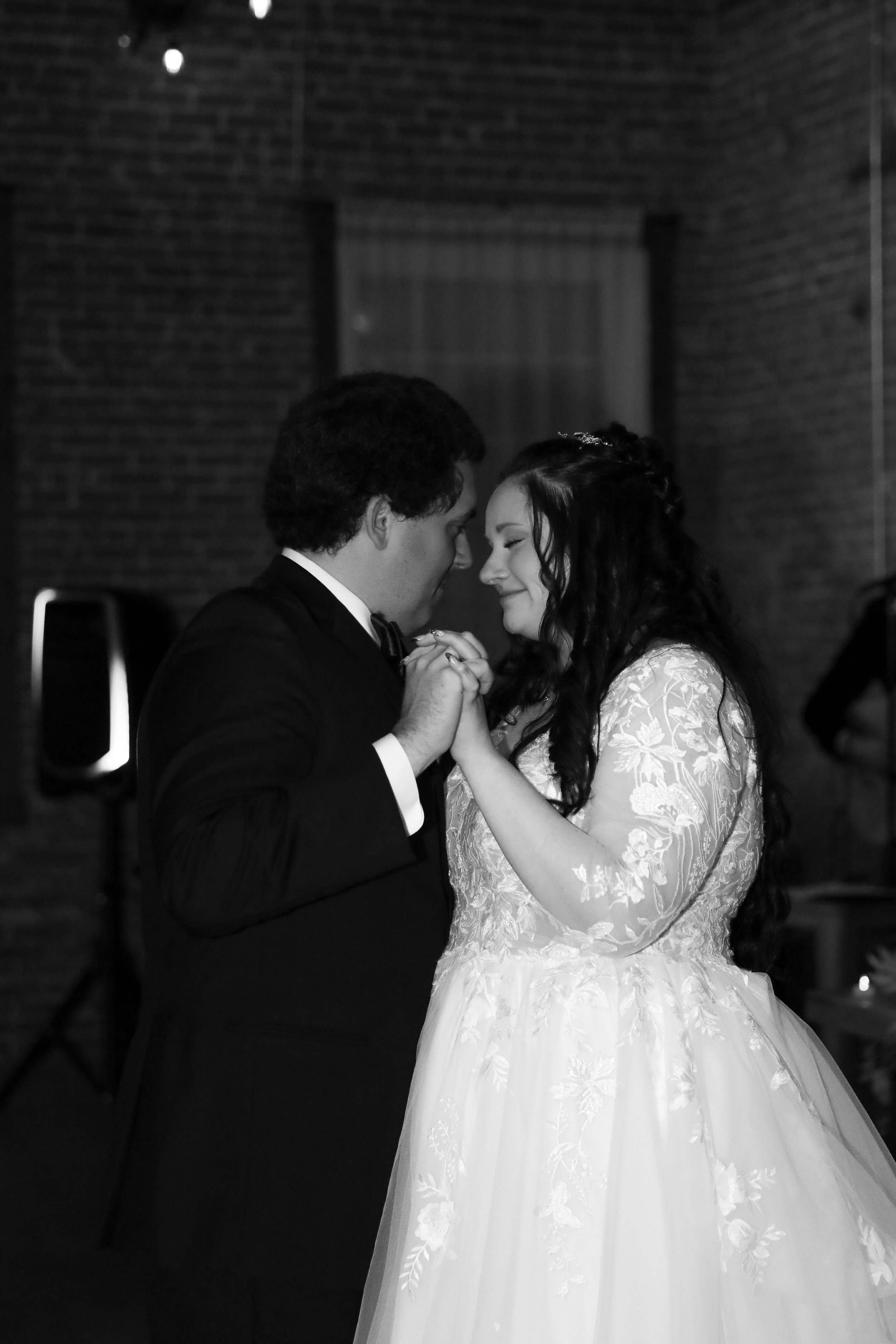 A black and white photo of a bride and groom dancing closely together, holding hands and smiling with their foreheads touching at a wedding reception.