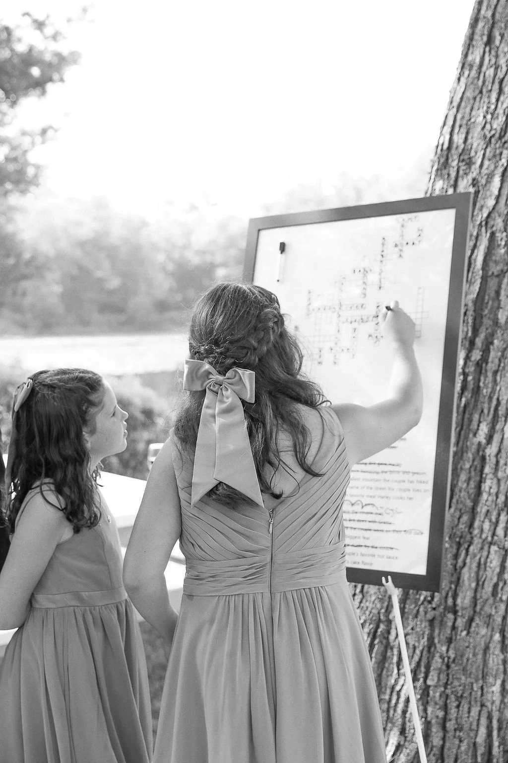 A woman with a large bow in her hair and in a sleeveless dress writes on a whiteboard with a marker, while a young girl in a similar dress watches. They are outdoors next to a large tree.