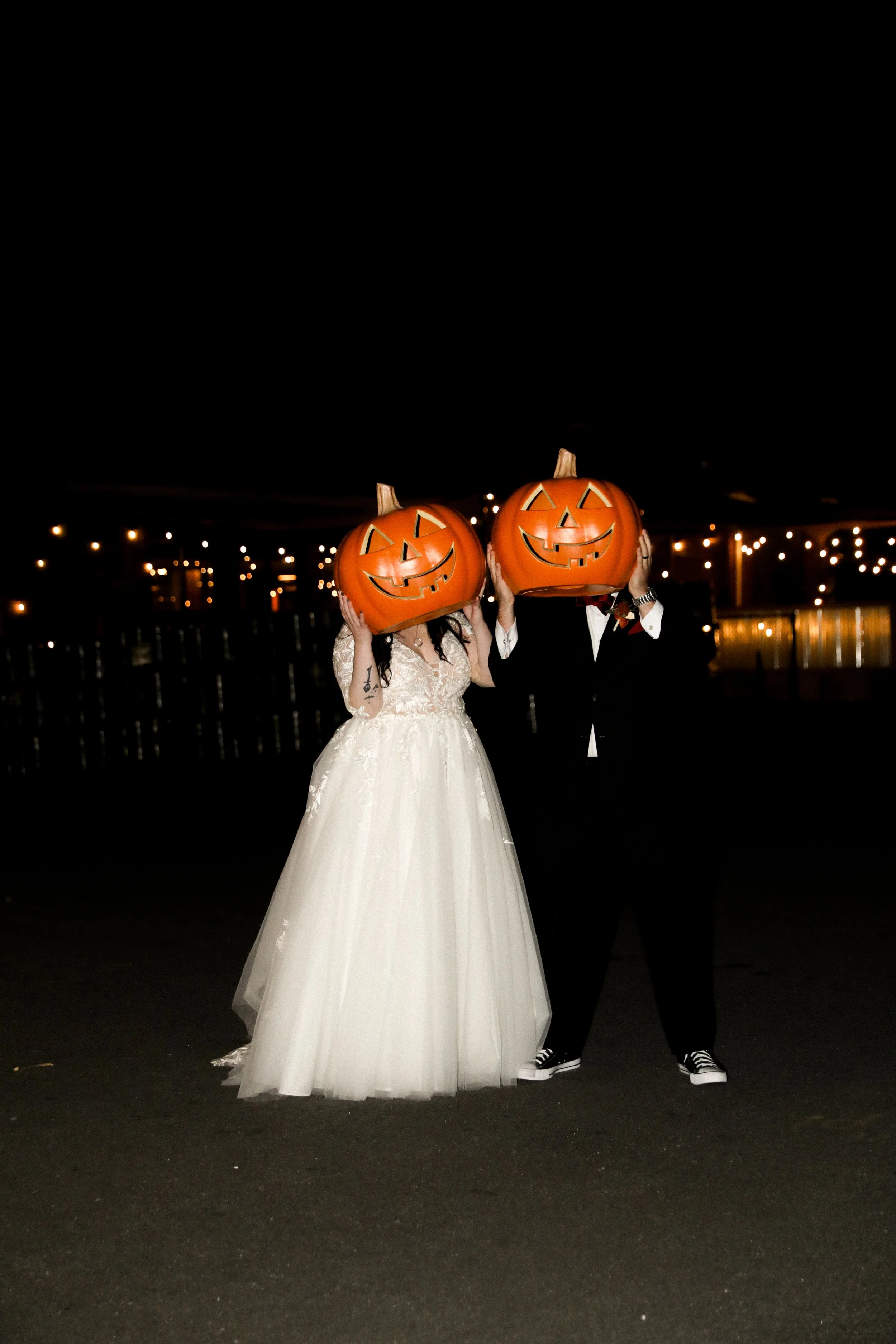 A bride and groom wearing wedding attire pose outdoors at night, each holding a large carved pumpkin with a jack-o'-lantern face in front of their faces.