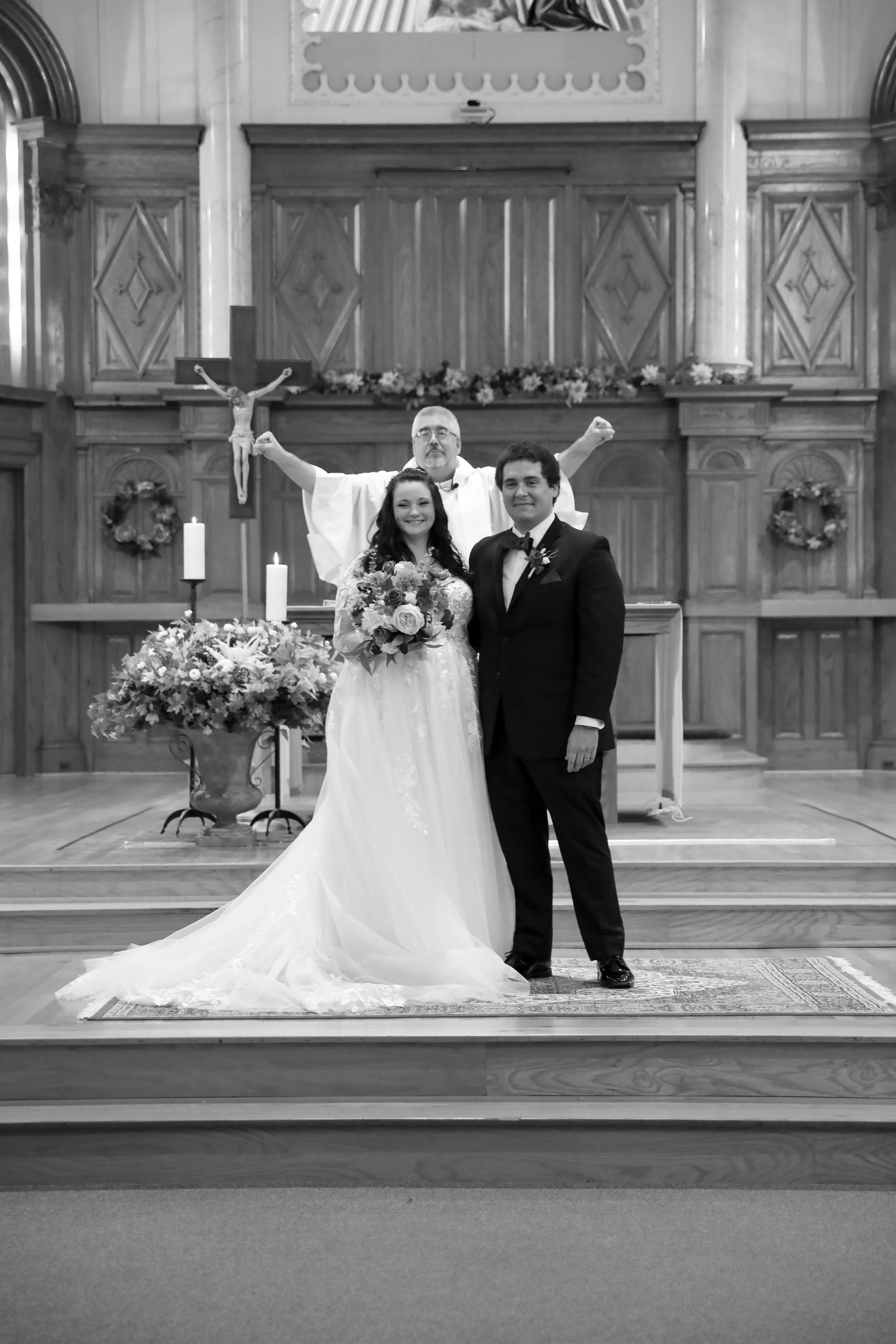 A wedding ceremony in a church with the bride and groom standing in front, the bride holding a bouquet, and the officiant in the background with raised arms.