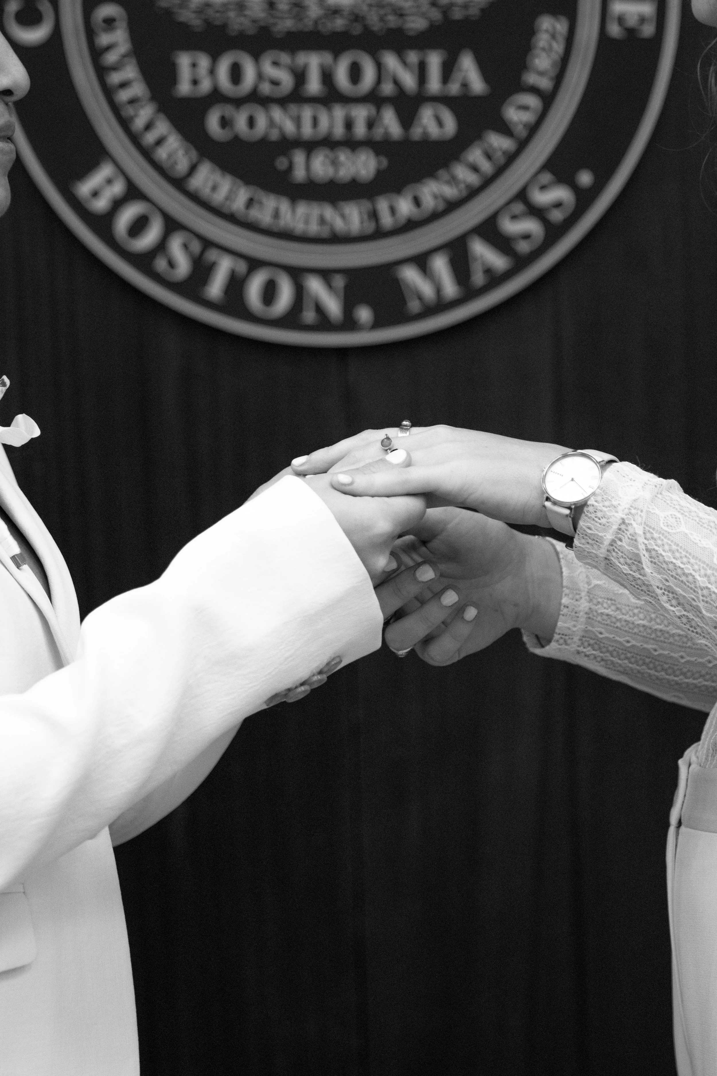 Two people exchanging rings during a wedding ceremony, with a Boston city emblem backdrop.
