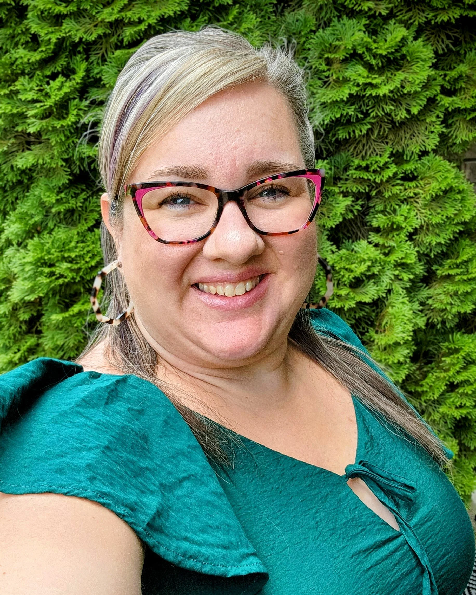Close-up of a smiling woman with blonde hair, wearing pink and tortoise shell glasses, standing in front of green foliage.