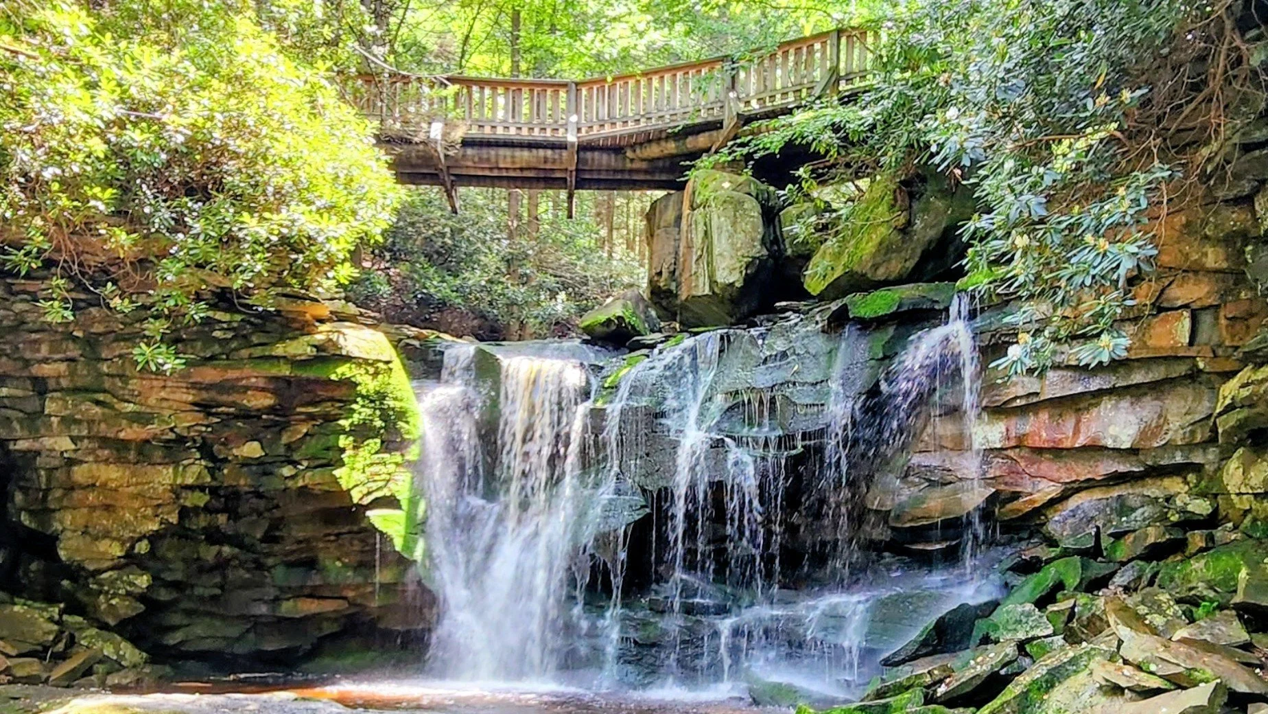 A wooden bridge over a multi-tiered waterfall surrounded by moss-covered rocks and lush green trees, sunlight filtering through the canopy.