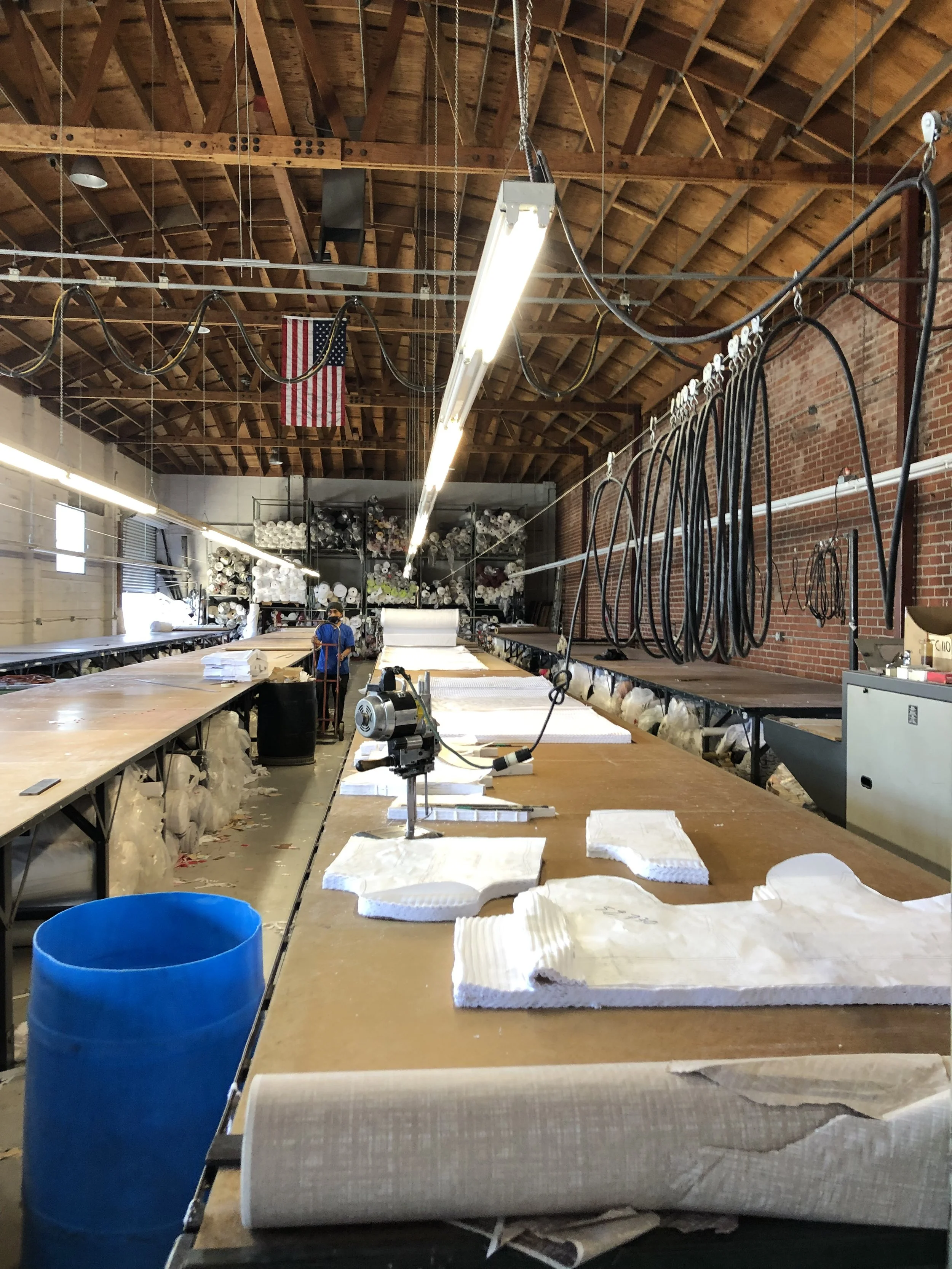 A large workshop with a high, wooden ceiling and brick walls contains work tables with foam molds, a worker in the distance, and shelves filled with rolled materials. Overhead lighting and coiled cables are hanging from the ceiling.