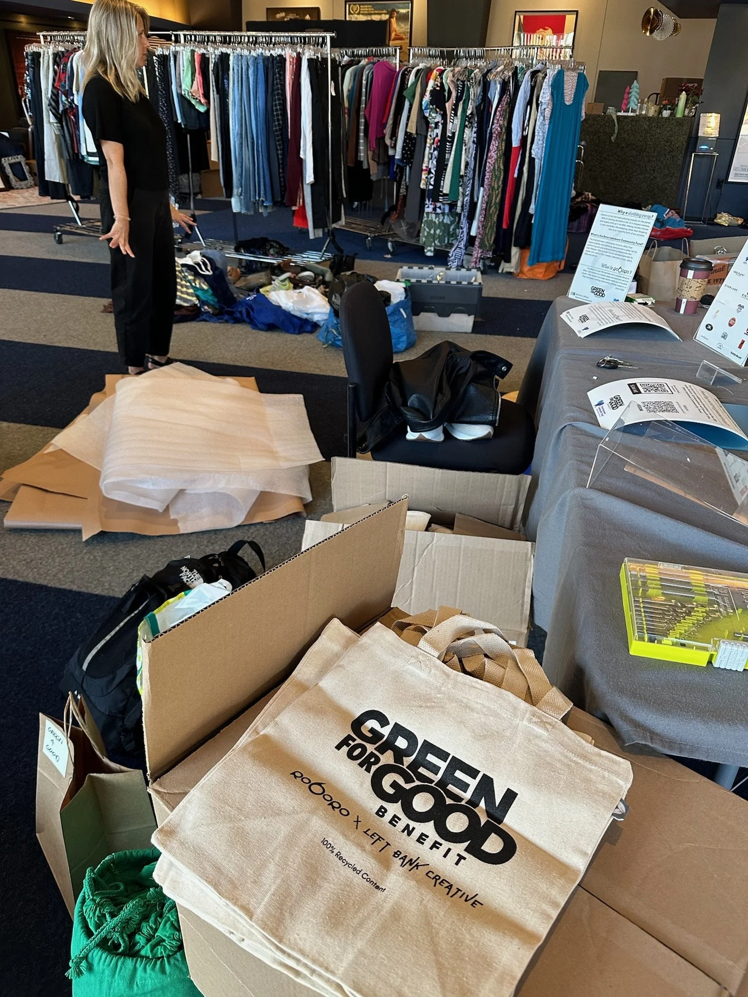 A woman shopping for clothes at a thrift store or charity sale, with racks of clothing in the background and various items and boxes on tables and the floor.