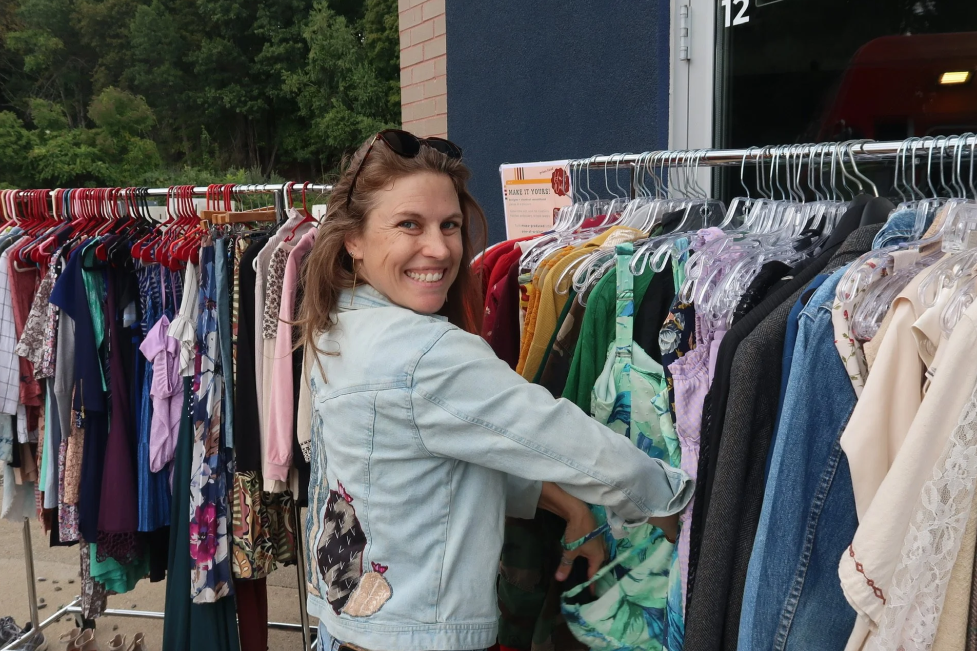 Smiling woman shopping for clothes at an outdoor thrift store, browsing through a rack of colorful garments.