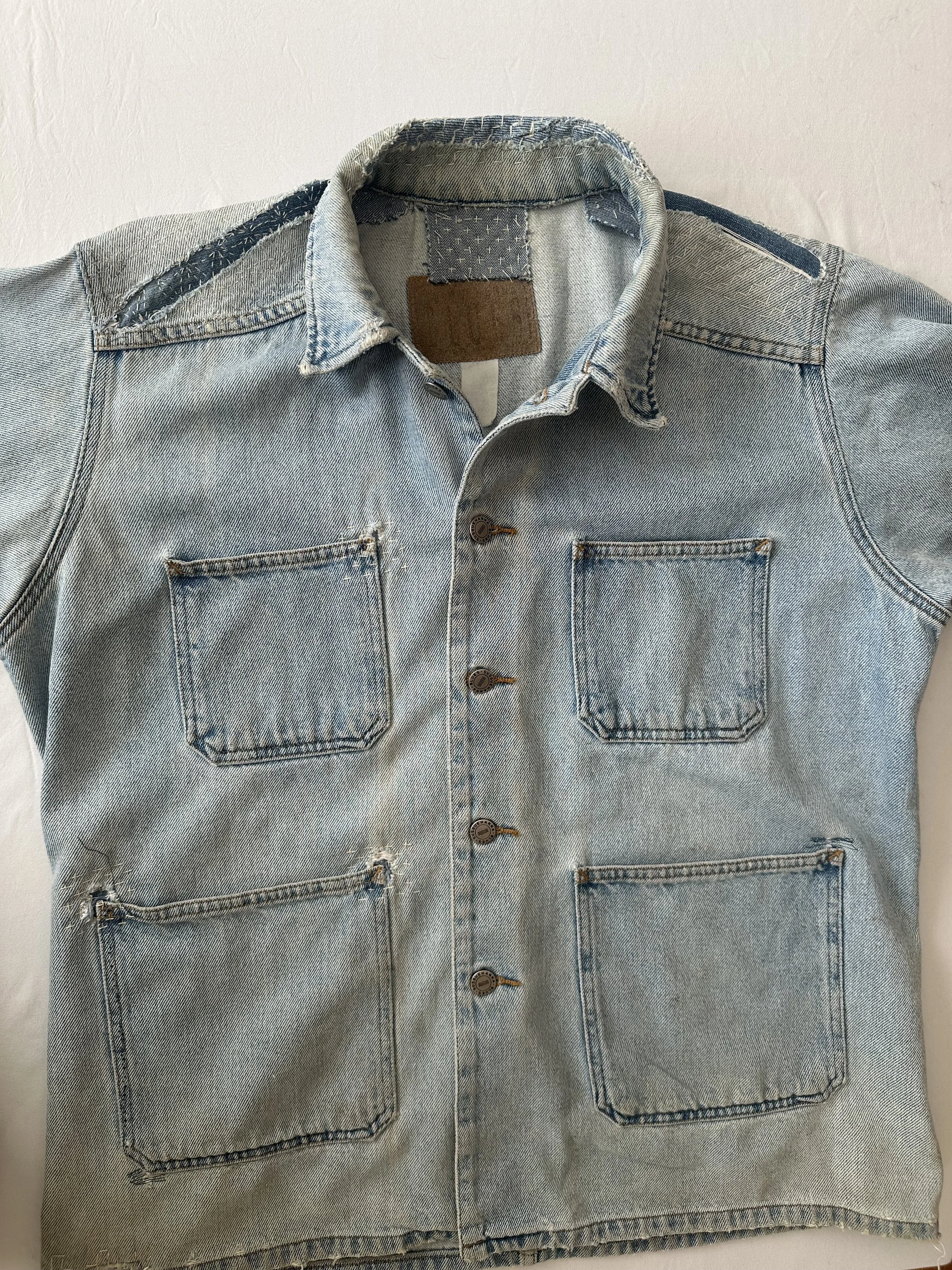 A light blue denim shirt with four front pockets, button closures, and a collar, laid flat on a surface.