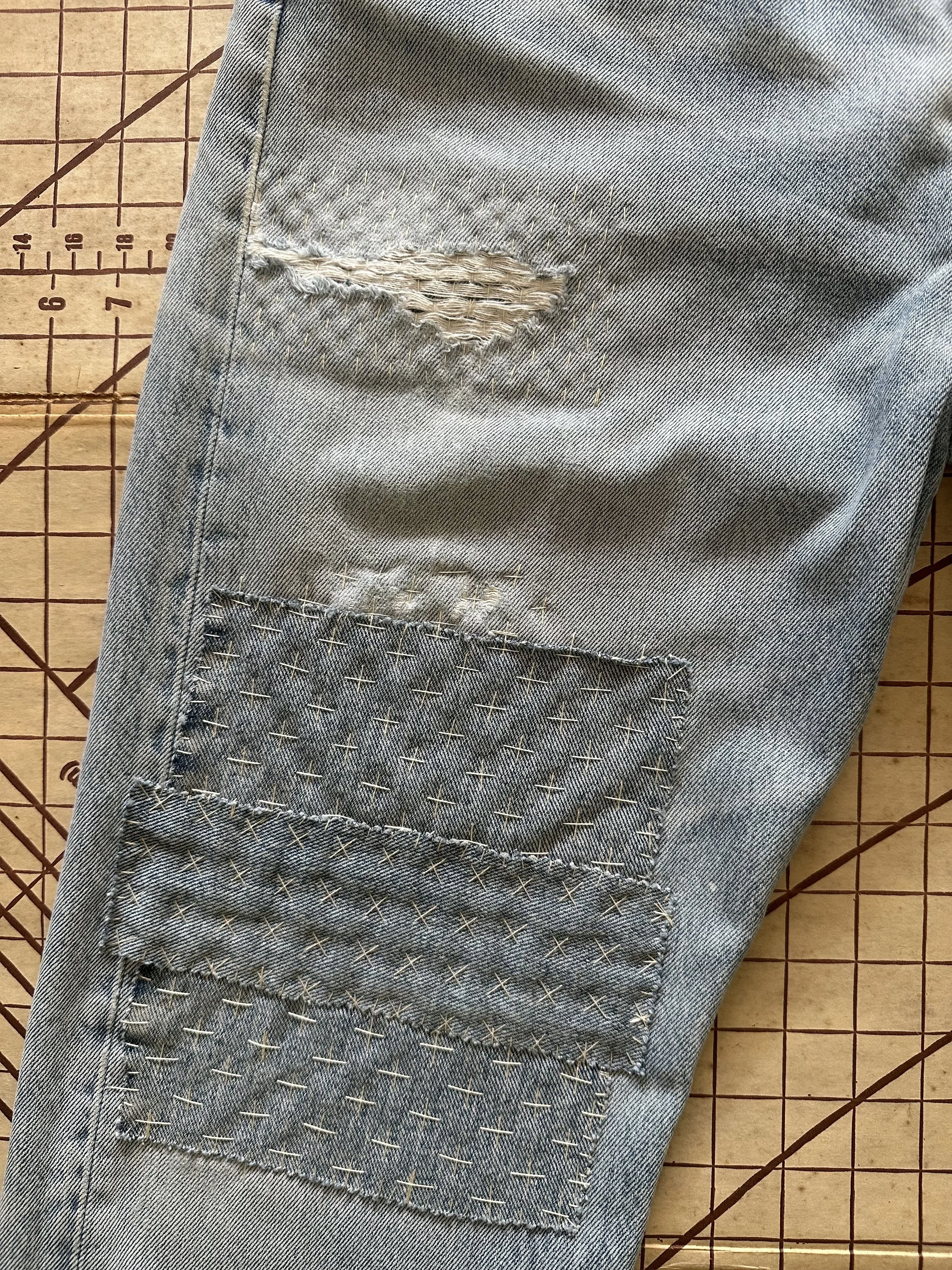 Close-up of a light blue denim pant with a ripped patch and stitched patches on the knee area, on a wooden grid cutting mat.