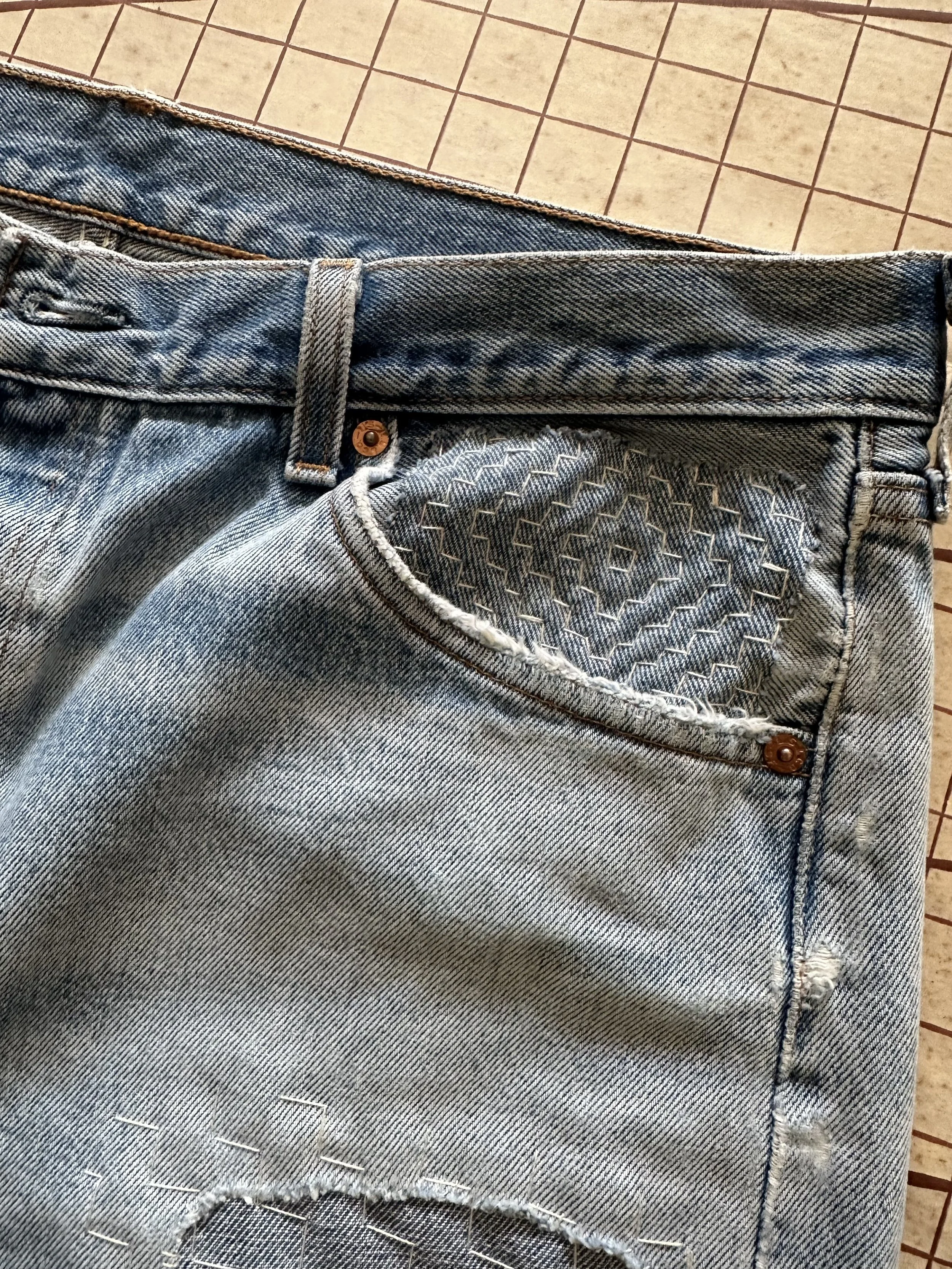 Close-up of a pair of light blue distressed denim jeans with a textured pocket detail, belt loop, and copper rivets, on a tiled floor.