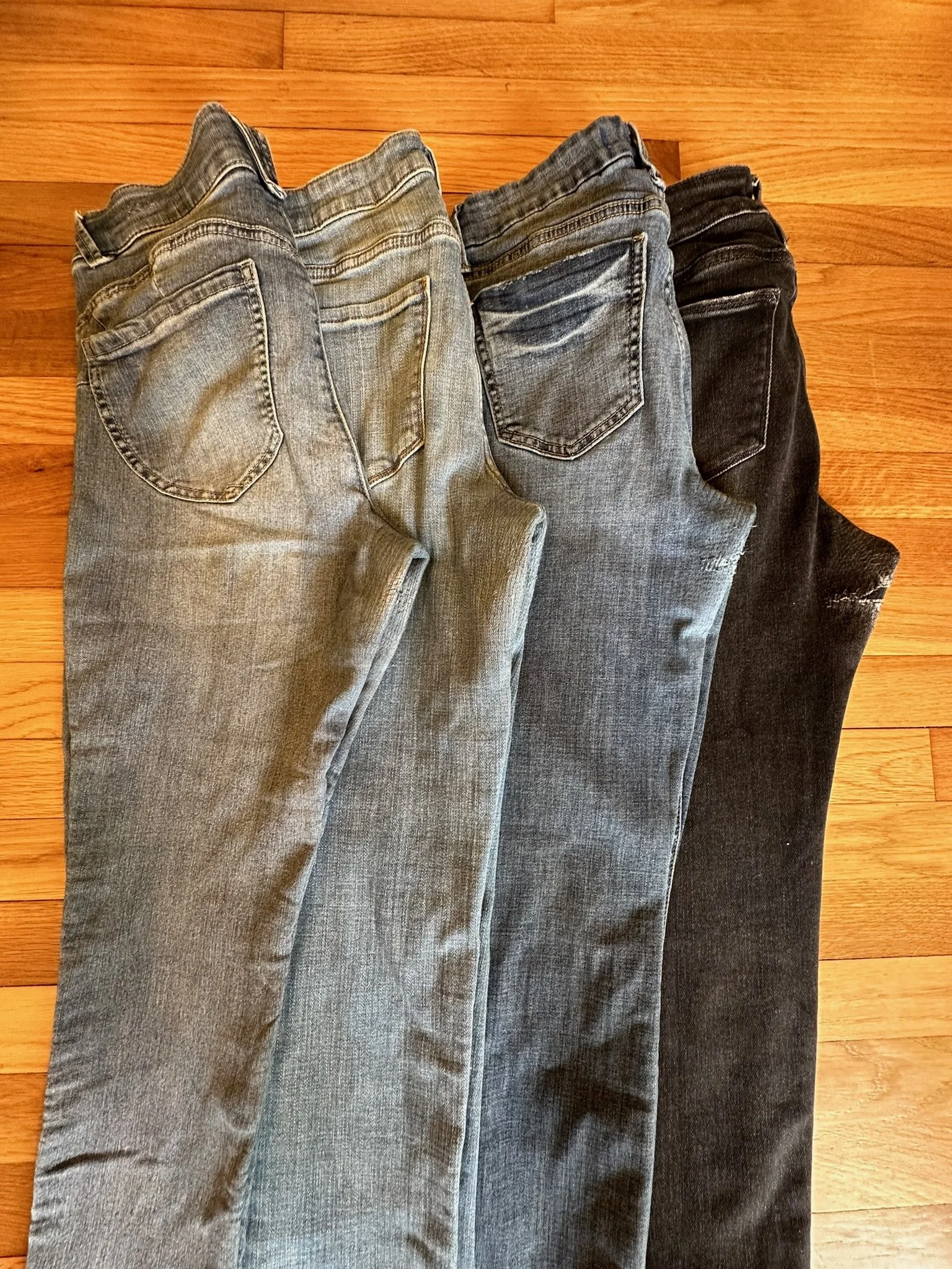 Three pairs of jeans laid flat on a hardwood floor, arranged in a row from lightest to darkest