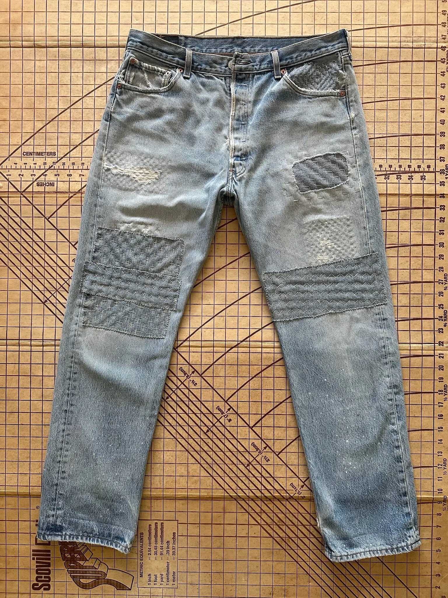 Gray distressed jeans with patches on the knees and thighs, laid flat on a wooden cutting mat with measurement markings.