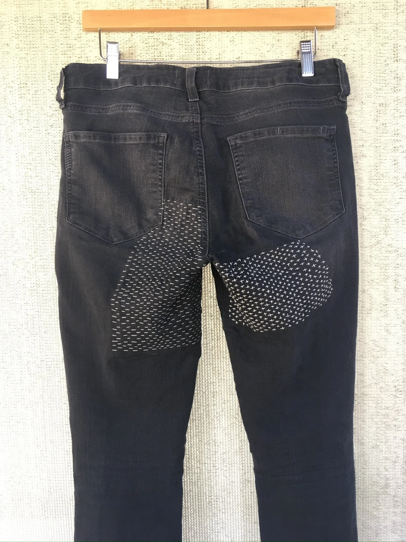 Back view of black denim jeans with circular patterned patches on the back of the knees, hanging on a hook against a textured wall.