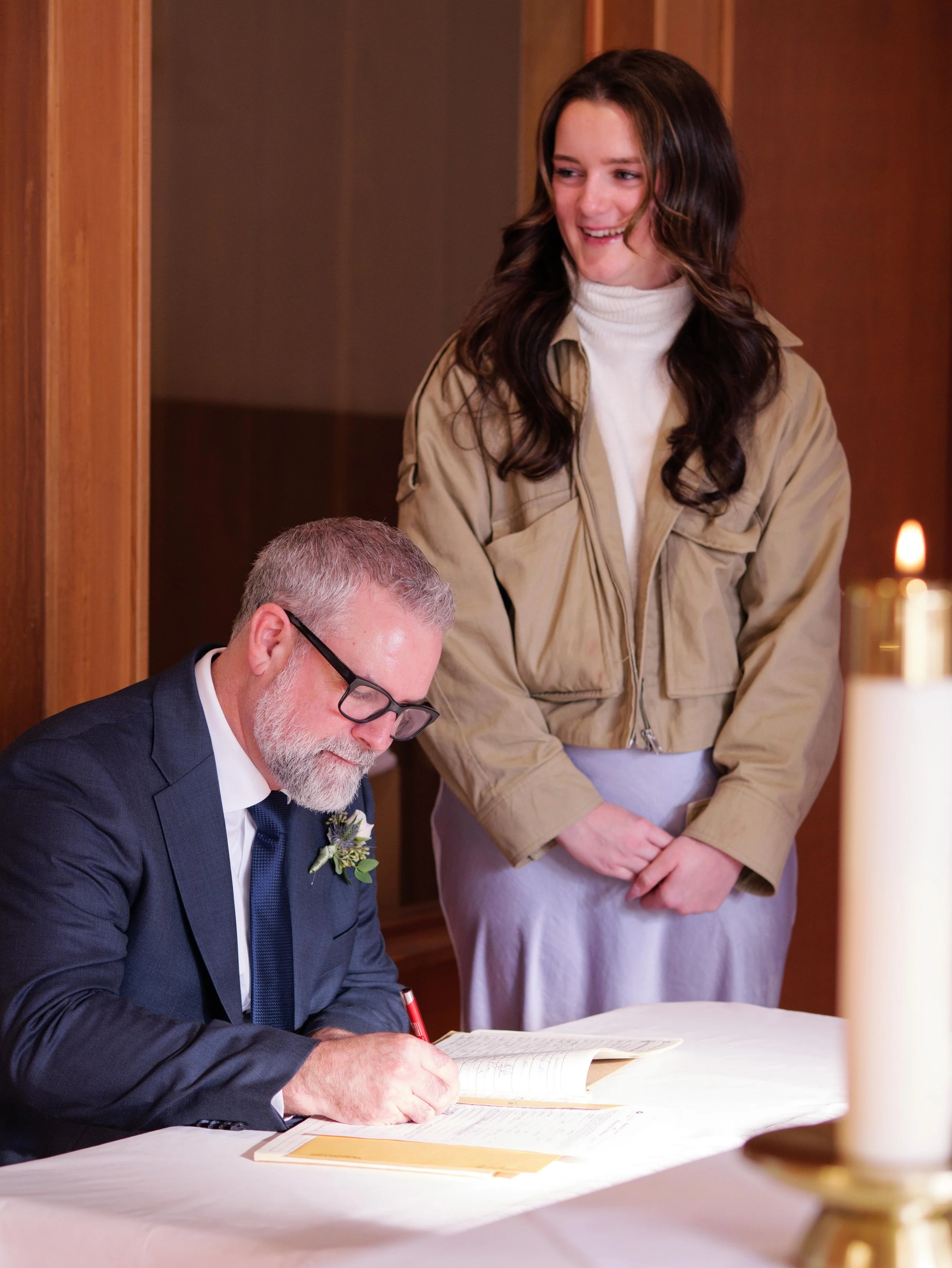 A man in a suit and glasses is signing a book or document at a table, while a woman stands beside him, leaning slightly and smiling, in what appears to be a formal setting with a lit candle in the foreground.