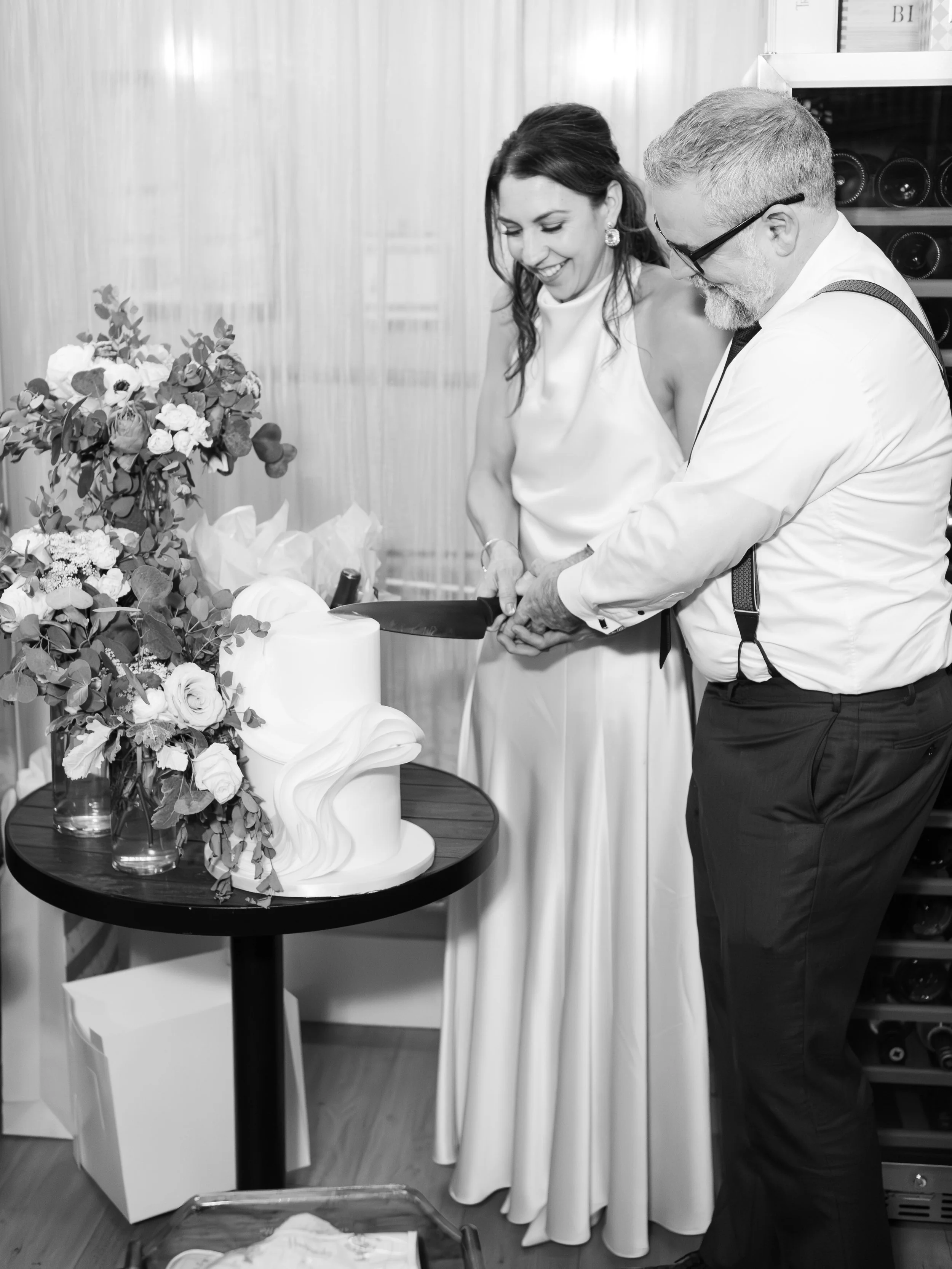 A man and woman cutting a wedding cake together at a wedding reception, surrounded by flower arrangements.