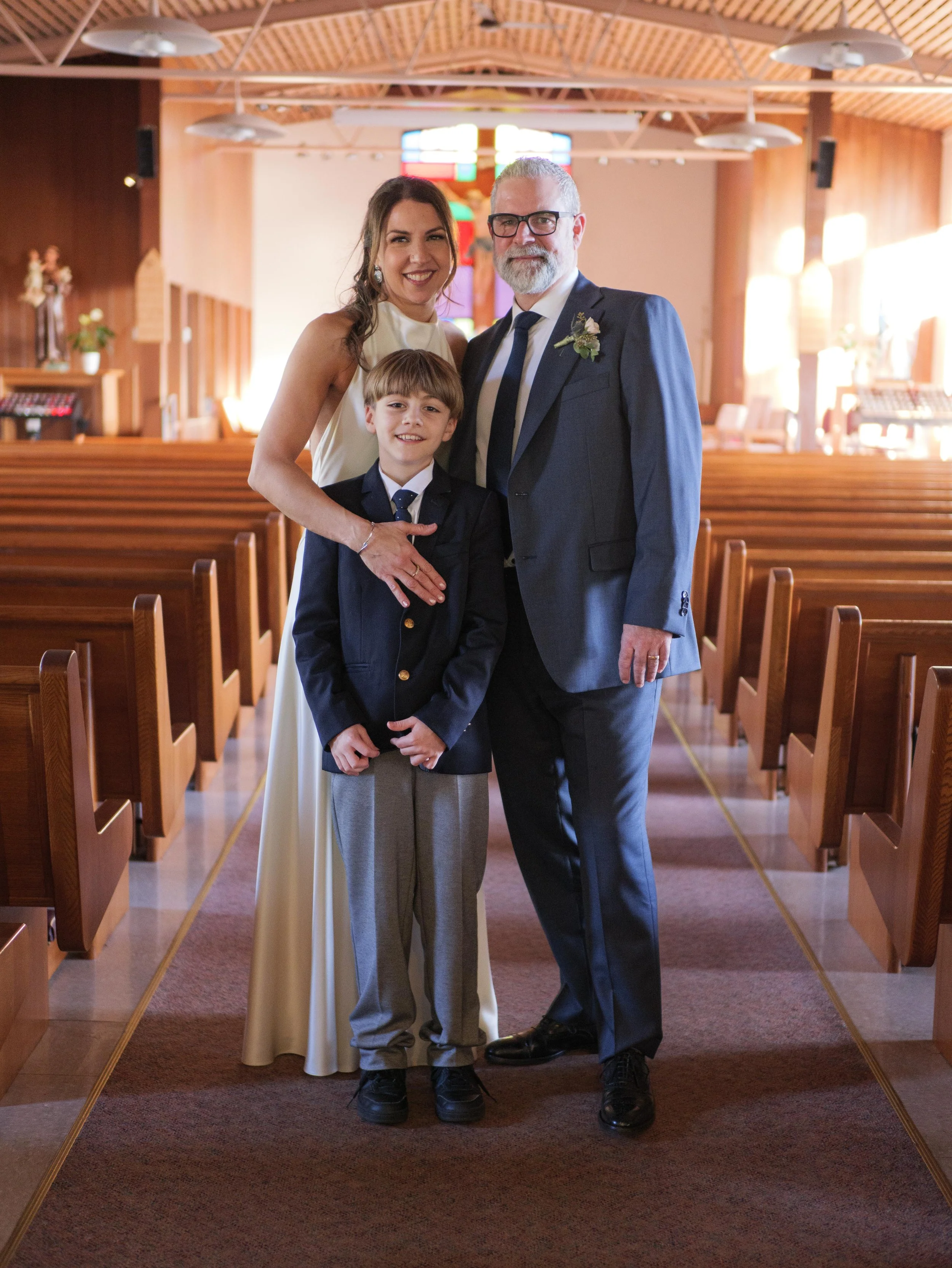 A family of three standing inside a church, smiling at the camera. The woman is in a cream dress, the man is in a suit with a boutonniere, and a young boy is dressed in a suit jacket and tie. The church has wooden pews and stained glass windows.
