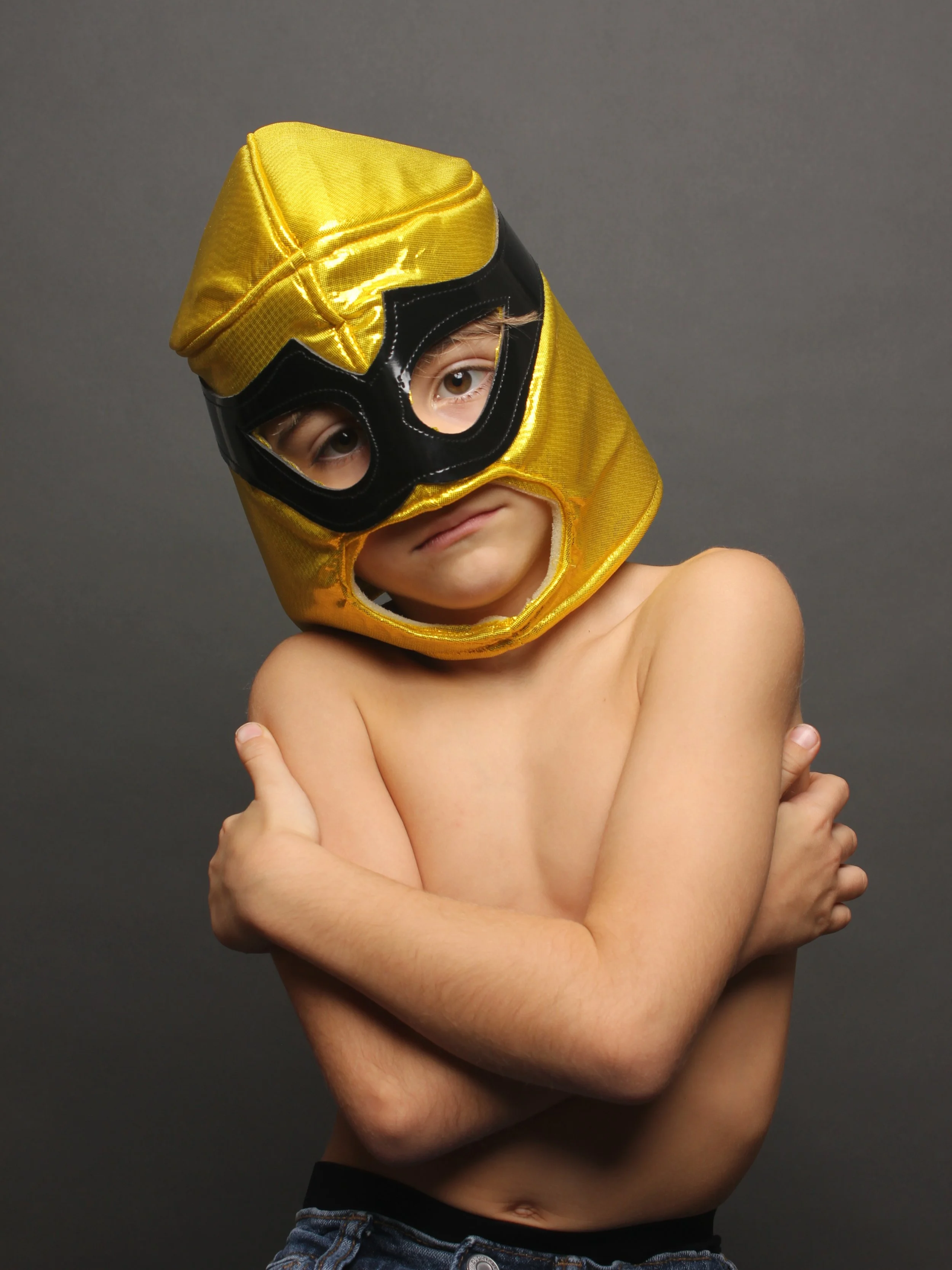 A shirtless boy wearing a gold and black superhero mask, crossing his arms with a serious expression against a plain gray background.