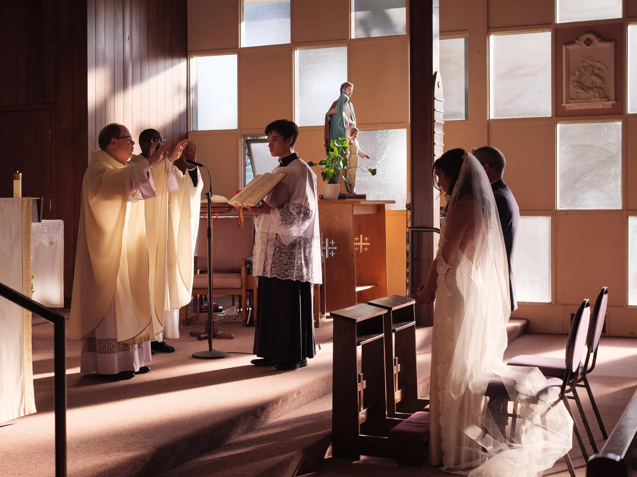 A Catholic wedding ceremony takes place inside a church with a priest, a minister, and a bride and groom kneeling in prayer. The priest and minister are wearing cream-colored robes, and the bride is in a white wedding gown with a veil.