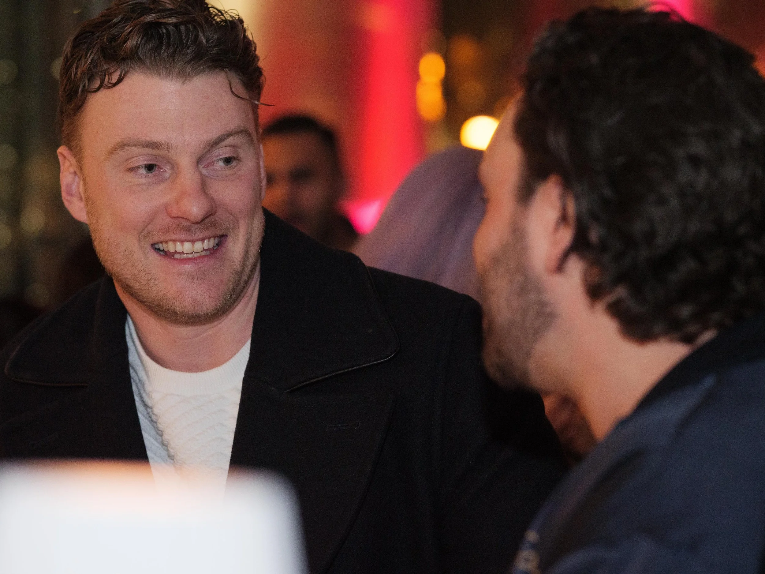 Two men engaged in a conversation at a social event, smiling and appearing friendly, with warm lighting and blurred people in the background.