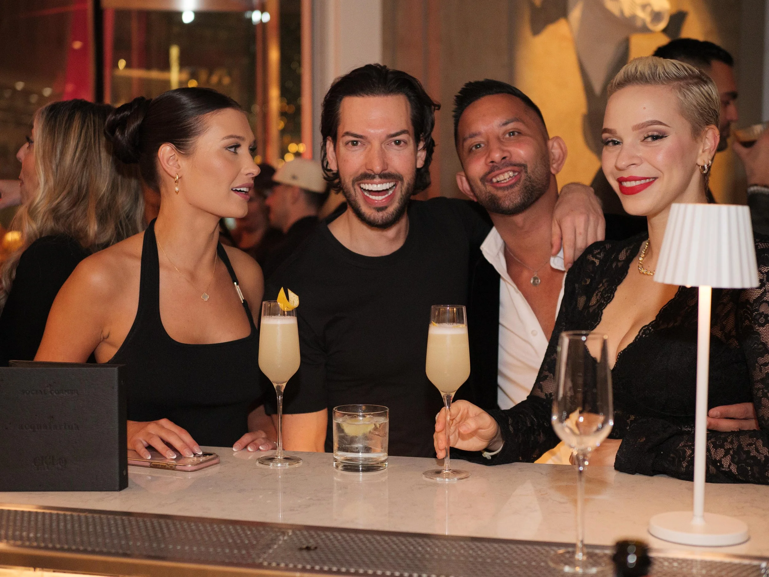 People smiling and enjoying drinks at a bar, with two women and two men in the foreground.