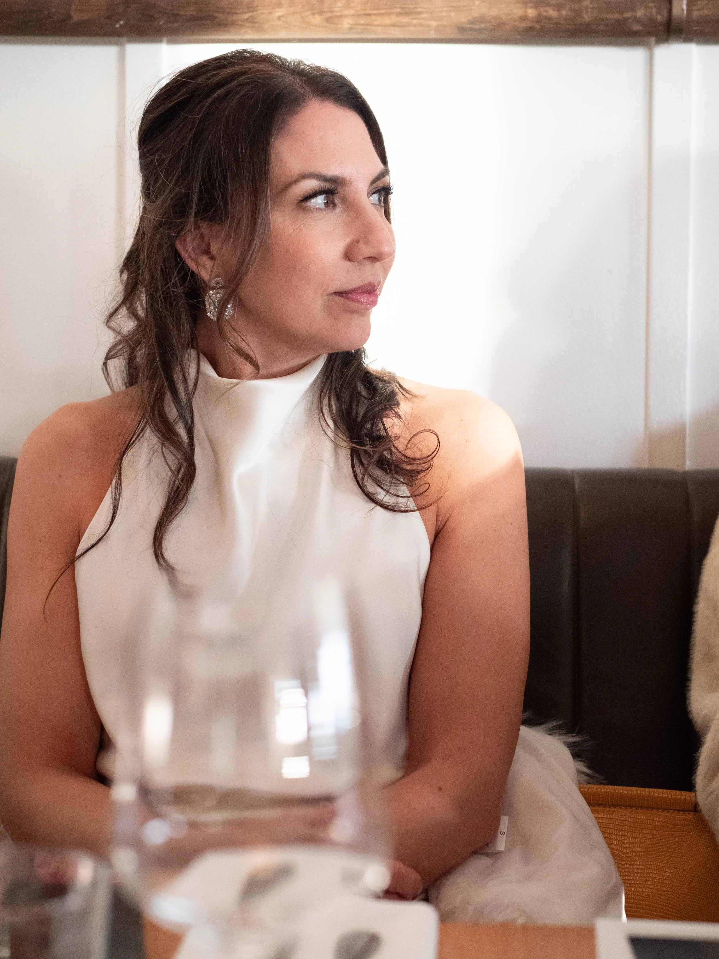 A woman with brown, wavy hair wearing a sleeveless white dress sitting at a table with a glass in front of her, looking to her left.