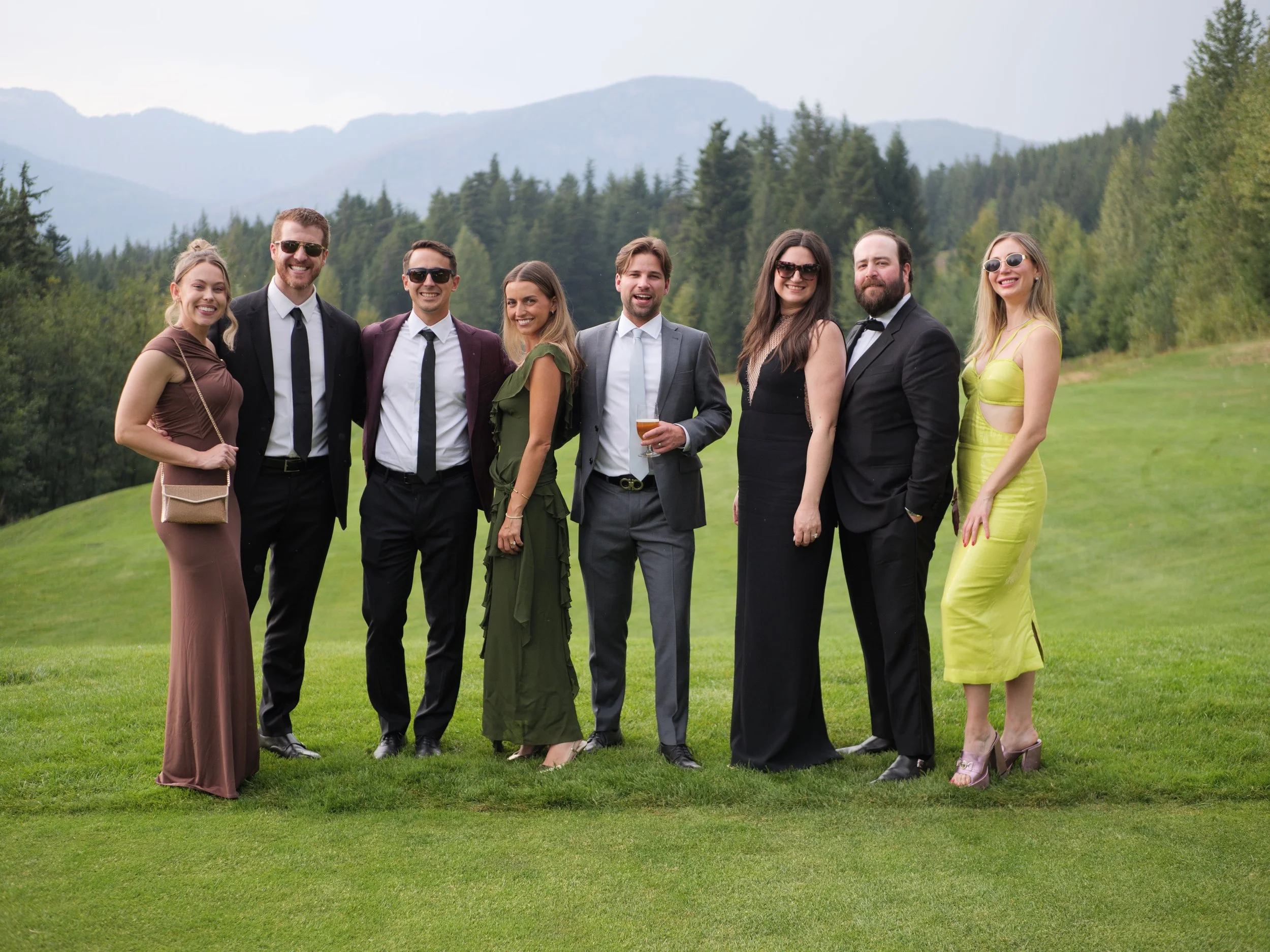 A group of nine adults dressed in formal and semi-formal attire, standing outdoors on a grassy area with mountains and trees in the background.