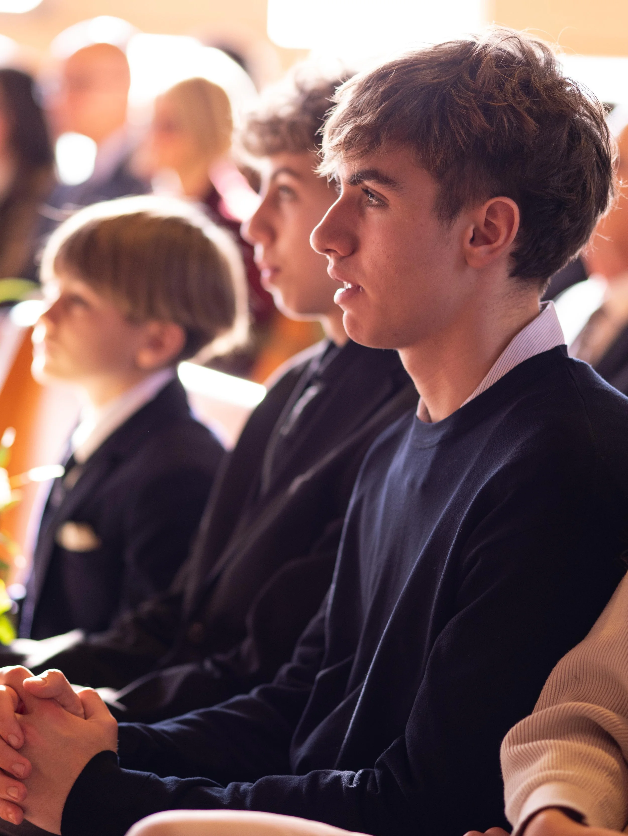 A group of teenagers sitting attentively during a formal event, dressed in dark clothing, with a focus on a young man with curly hair in the foreground.