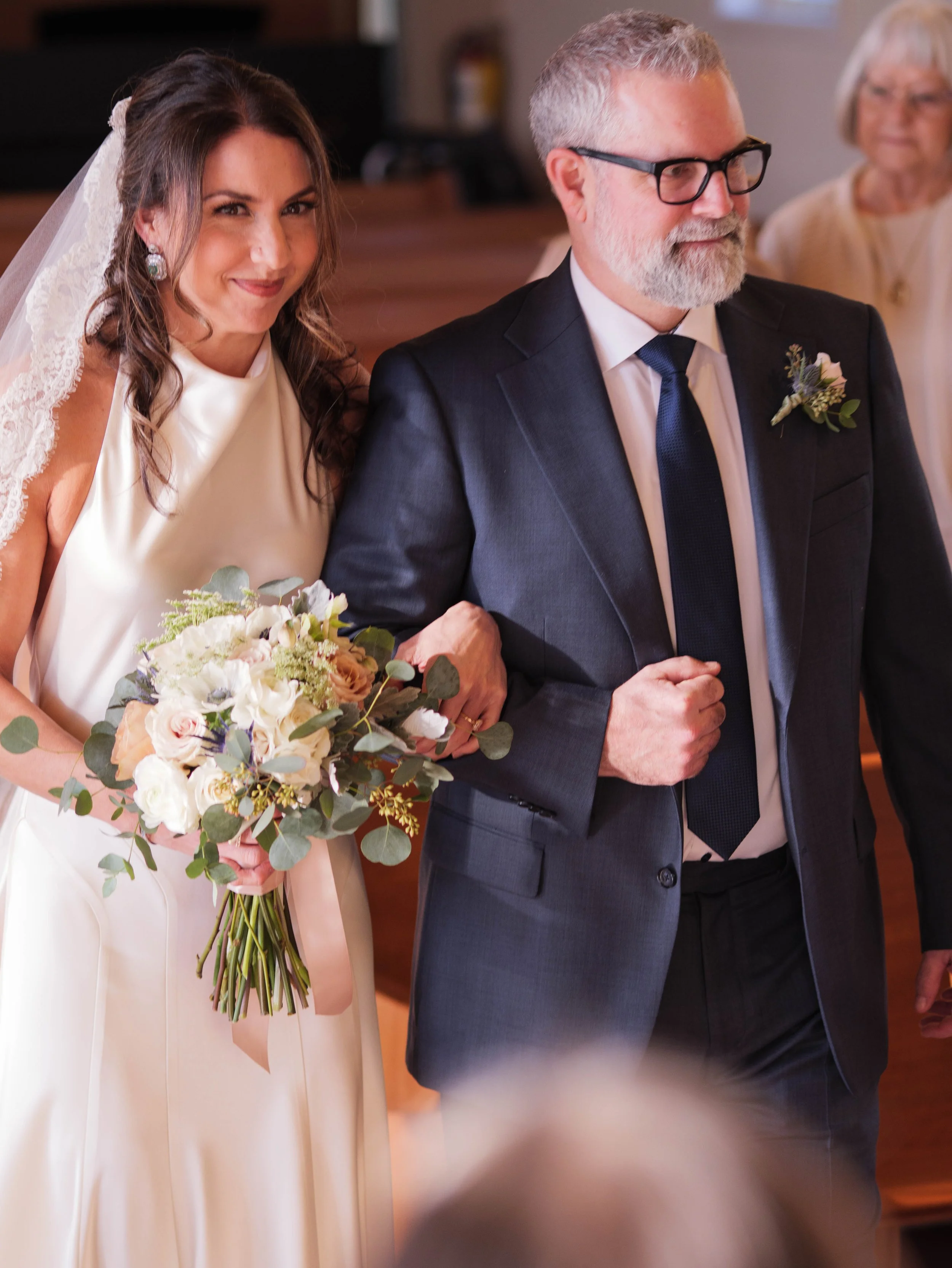 A bride in a cream dress holding a bouquet of white, blush, and green flowers, walking down the aisle with an older man in a navy suit and glasses, while an older woman looks on in the background.