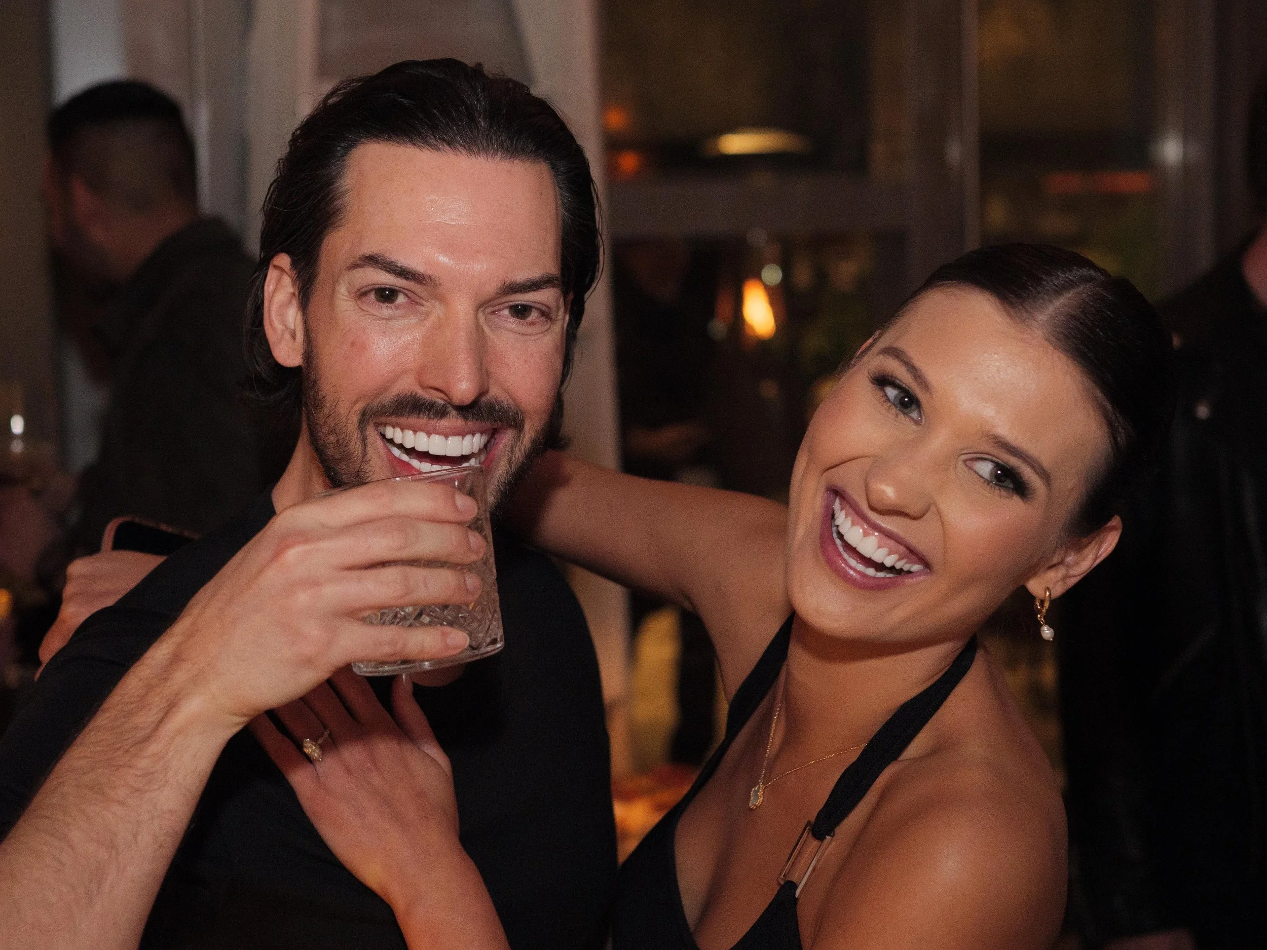 A man and woman smiling at a party, with the woman having her arm around the man. The man is holding a drink in his hand, and both appear to be happy and enjoying themselves.