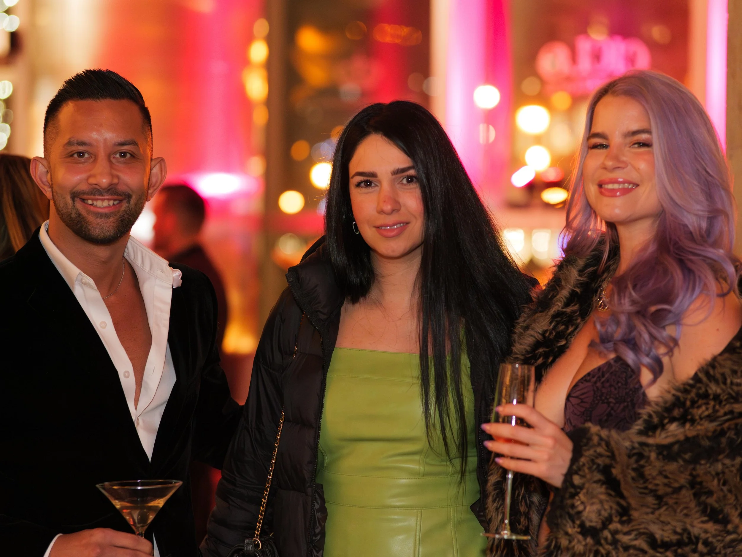 Three people, two women and one man, at a lively event with colorful blurred lights in the background. The man on the left is holding a martini glass, smiling. The woman in the middle has long black hair, wears a green dress, and has a neutral expres