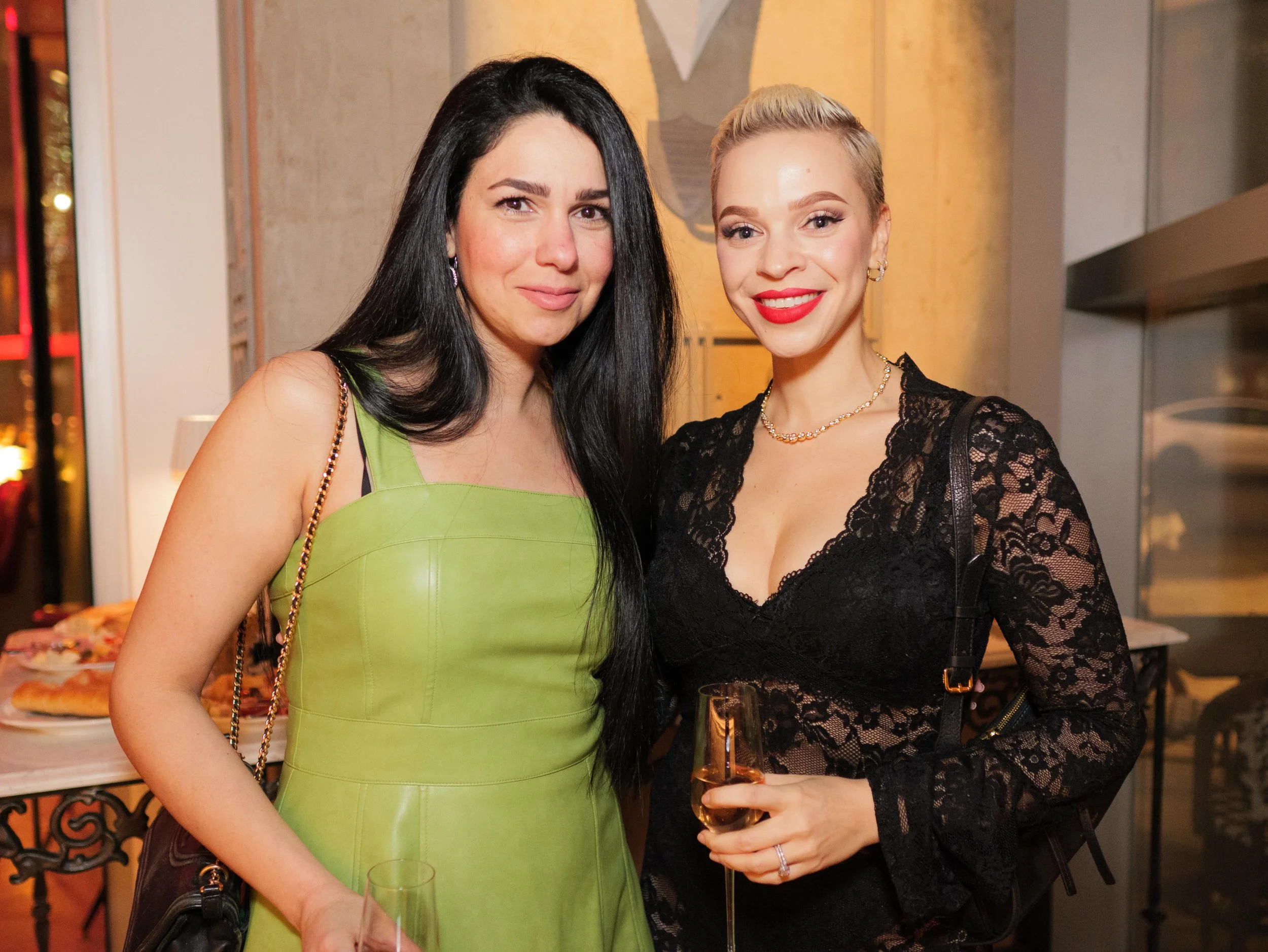 Two women at a social event, one in a green dress and the other in a black lace dress, holding glasses of champagne and smiling at the camera.