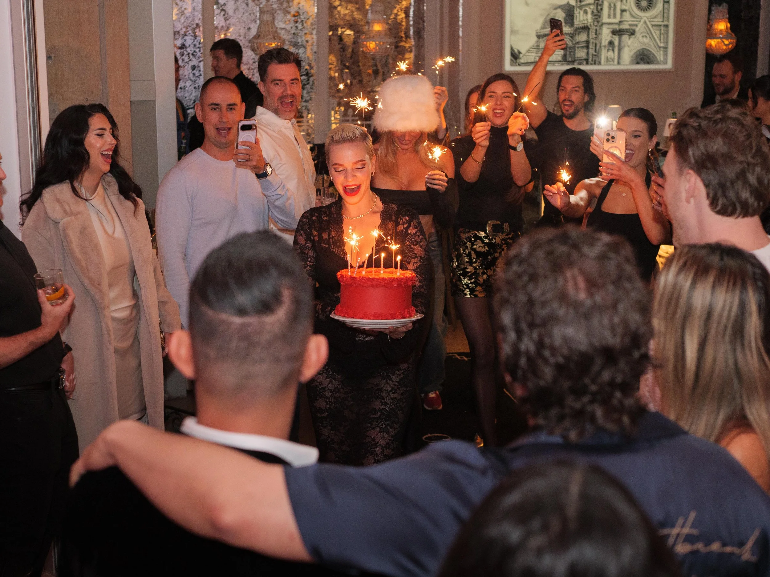 A woman holding a red birthday cake with lit candles, surrounded by smiling friends at a celebration, some capturing the moment with phones, in a warmly lit party setting.