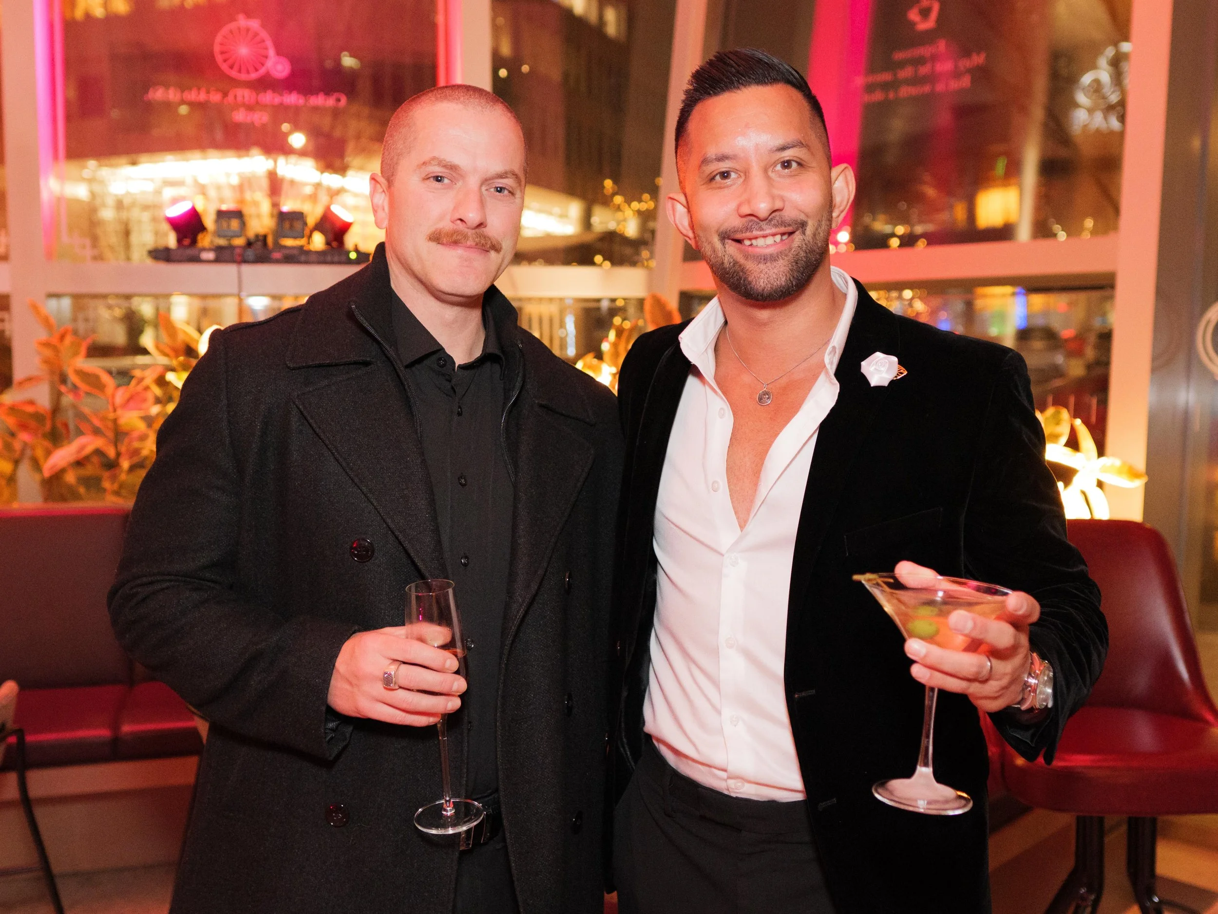 Two men dressed in formal attire at an indoor venue with city lights outside. One man with a mustache in a black coat holds a champagne glass, and the other man with a beard in a white shirt and black blazer holds a martini glass.