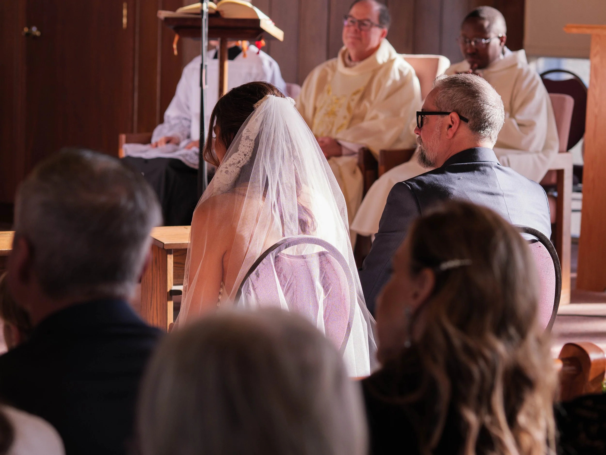 A bride and groom sit facing each other during a wedding ceremony in a church, surrounded by seated guests and officiants.
