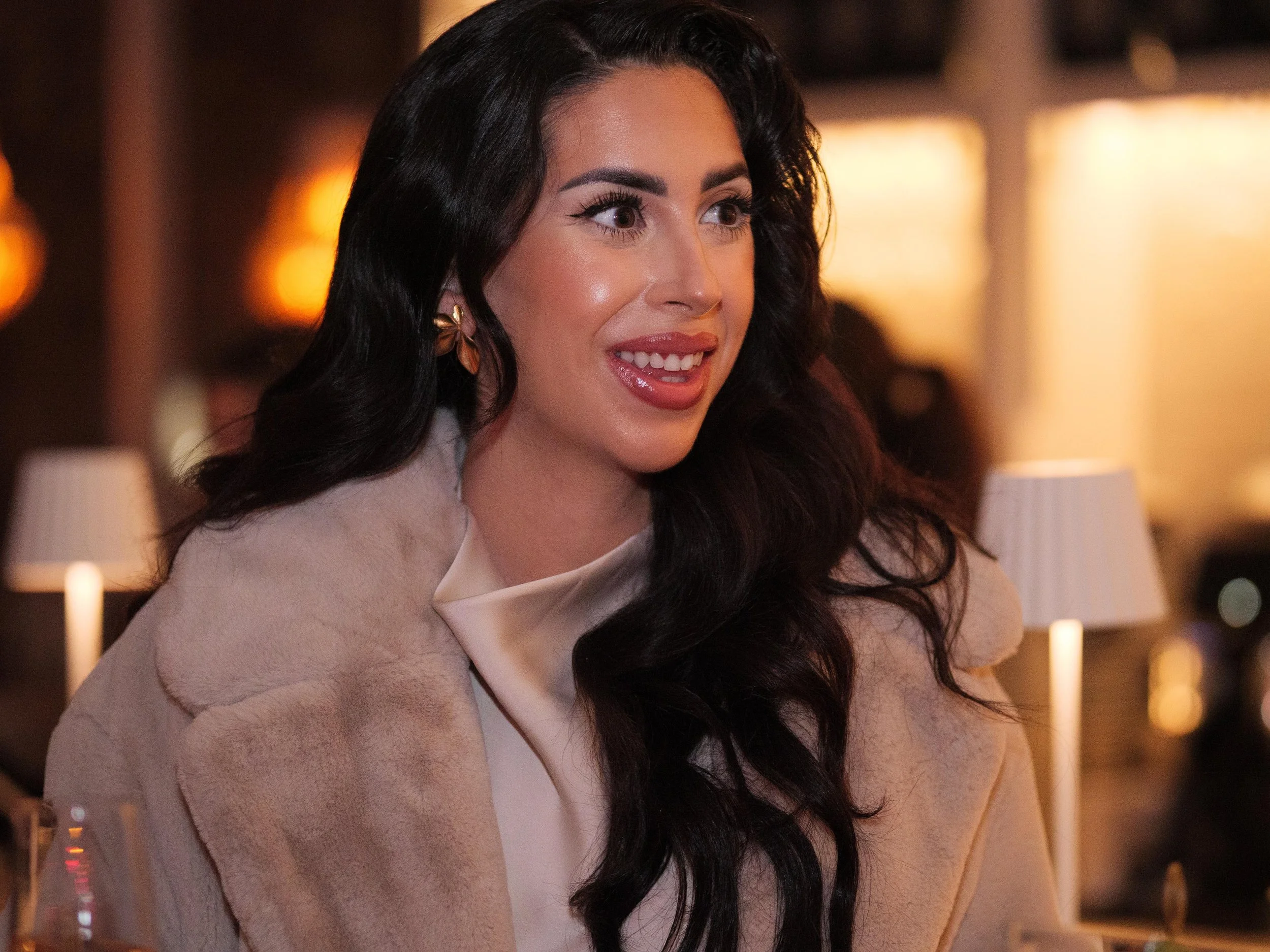 A woman with long dark hair, wearing makeup and a beige fur coat, smiling in a warm, dimly lit setting.