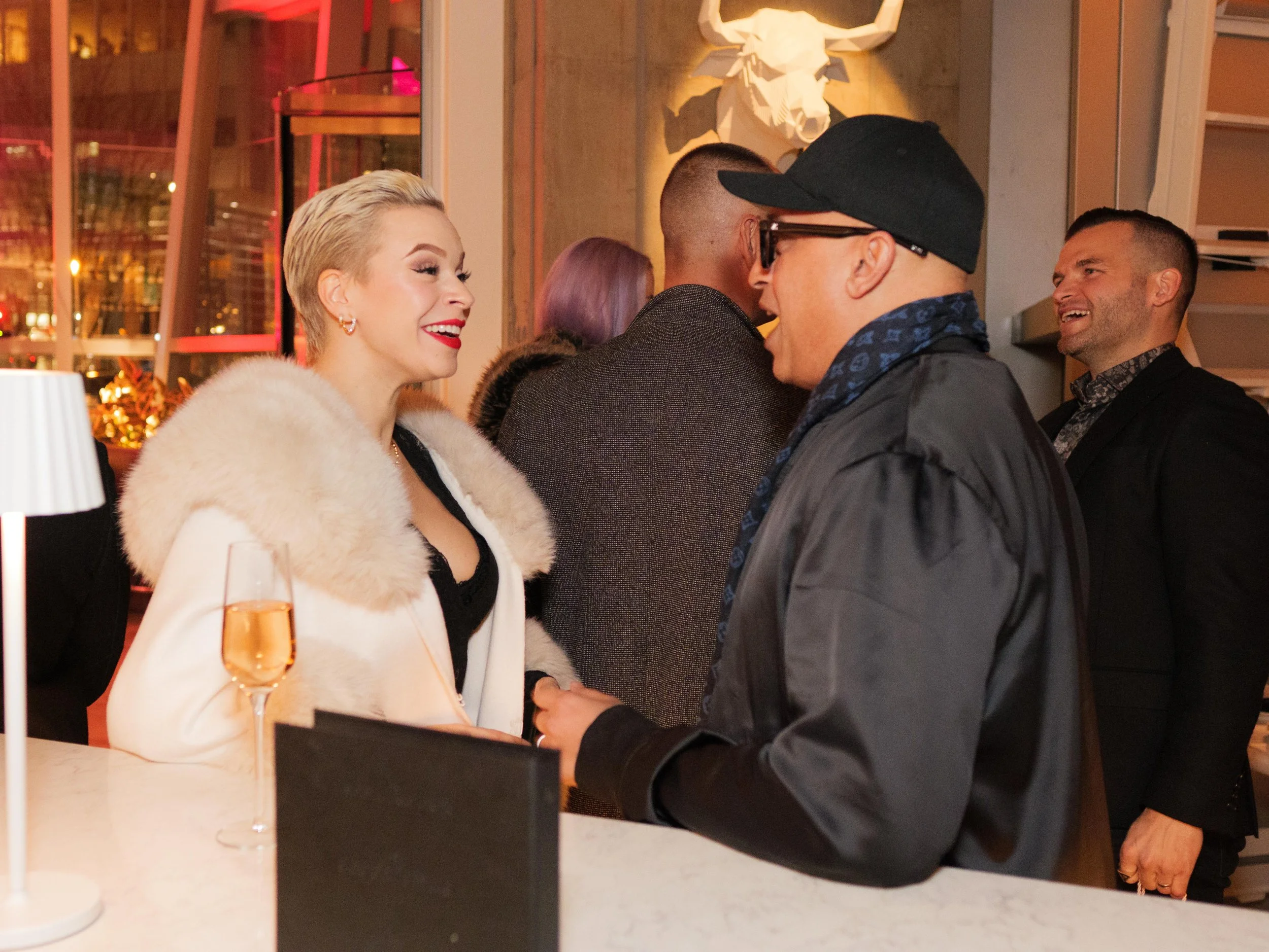 A woman with short blonde hair wearing a white fur coat and red lipstick is smiling and talking to a man wearing glasses, a black cap, and a dark jacket. They are at a bar or reception area with a champagne glass and a small black sign on the counter