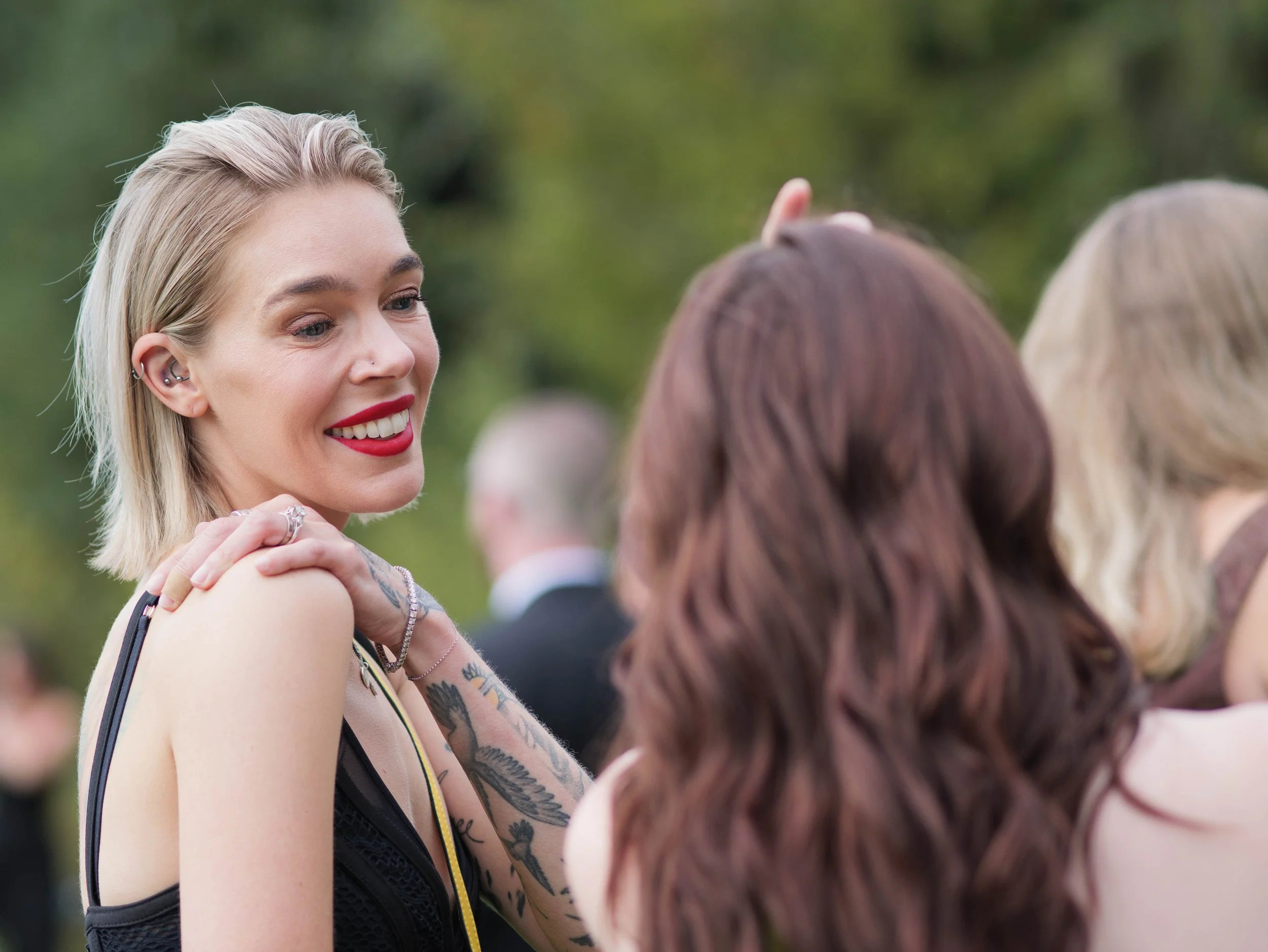 A blonde woman with tattoos smiling and talking to two women with brown and blonde hair at an outdoor gathering with trees in the background.